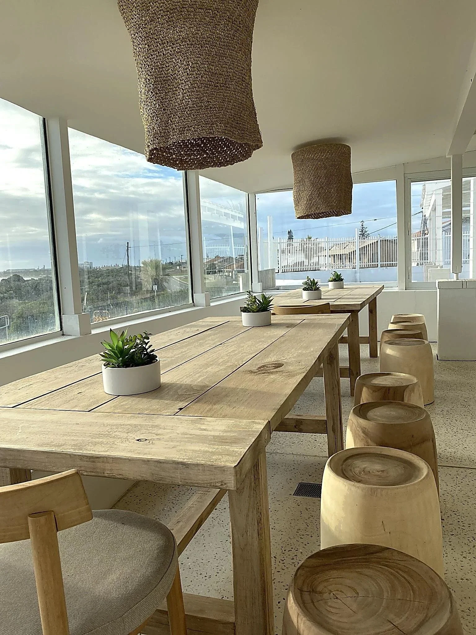 The Beach House Restaurant with large windows showing an outdoor view, a wooden table with small potted succulents, and hanging woven light fixtures.
