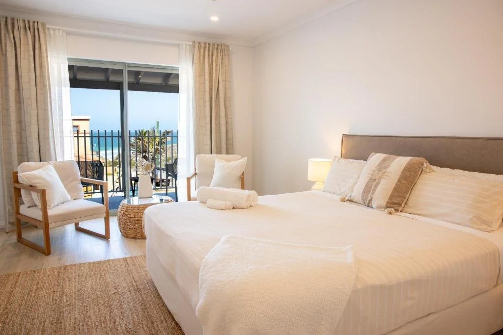 A bright hotel bedroom with a large bed, two pillows, a small table, two armchairs on a balcony, and ocean view through glass sliding doors.