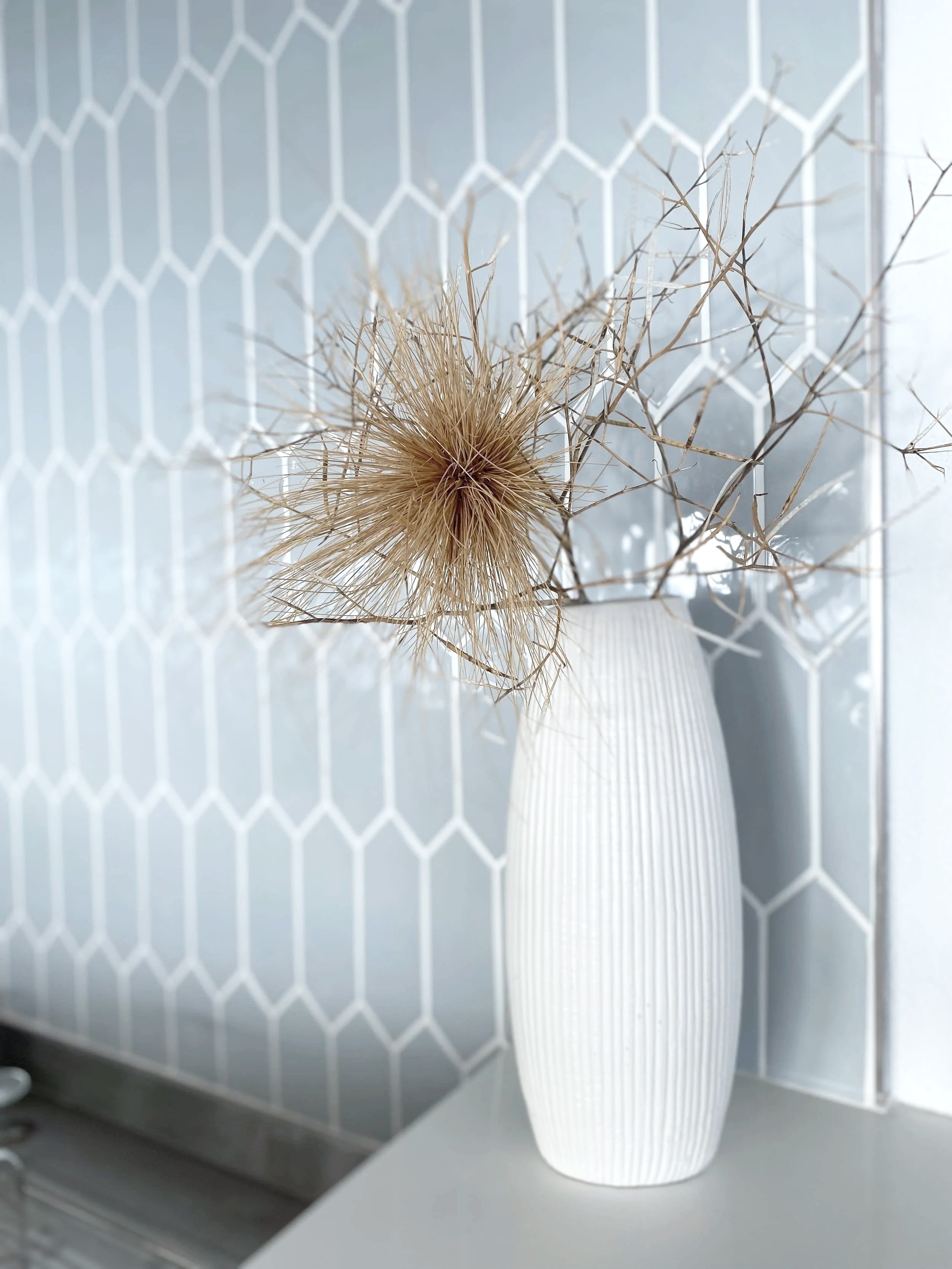 A white, vertically ribbed vase with dried, twig-like branches is placed on a white surface in front of a light gray wall with a hexagonal overlay pattern.