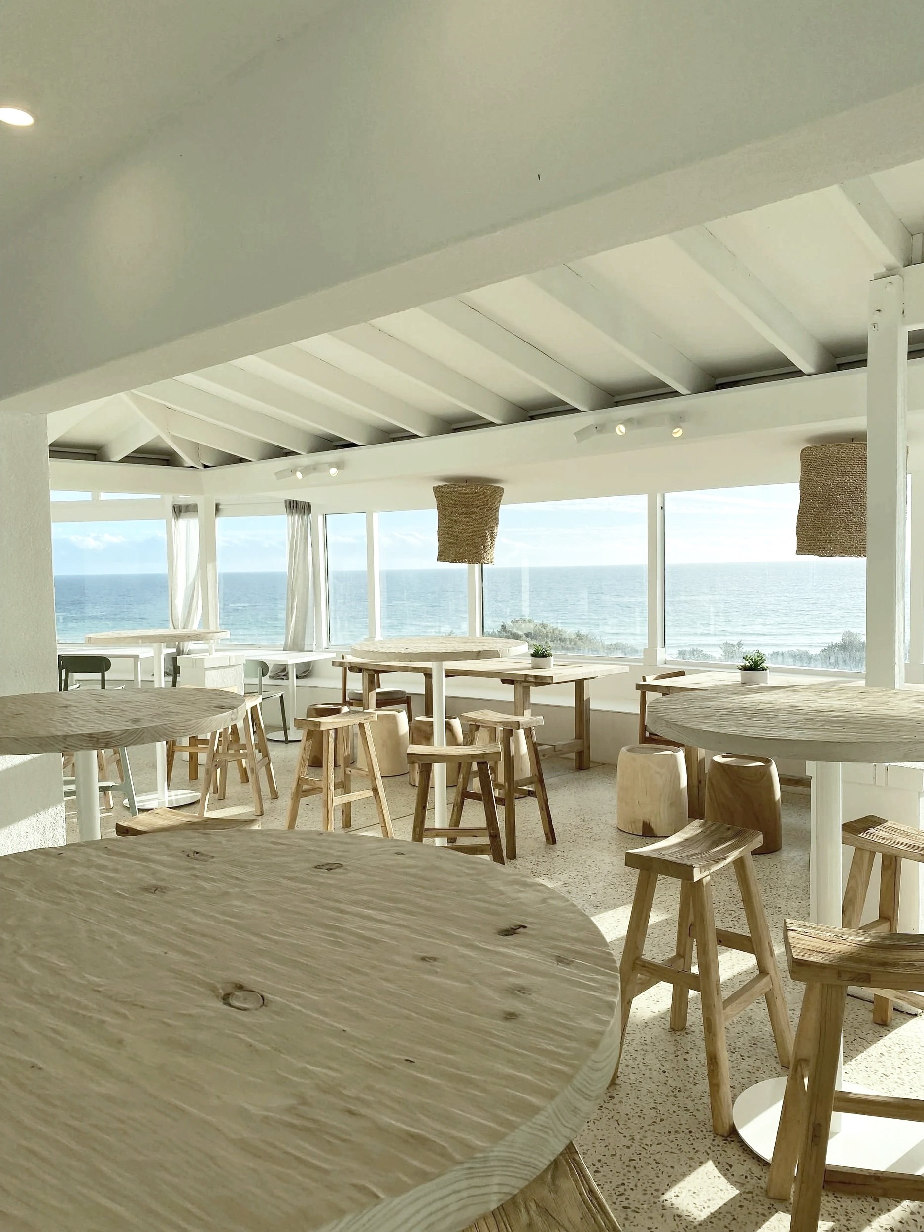 Interior of a bright seaside restaurant with wooden tables and stools, large windows showing the ocean, and natural lighting.
