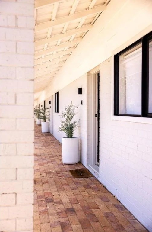 A white brick building with a covered porch, featuring potted plants and large windows.