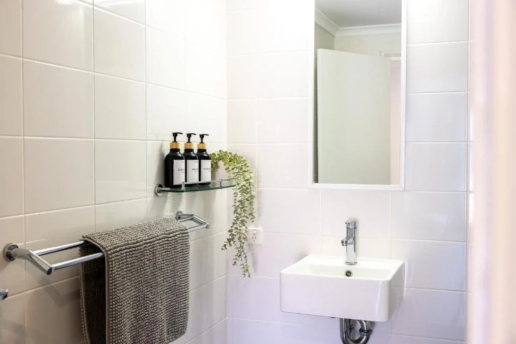 A small bathroom with white tiled walls, a white sink with a chrome faucet, a mirror above the sink, a glass shelf holding three black bottles of soap or lotion, a hanging green plant, a gray towel on a chrome towel rack, and a partially visible open