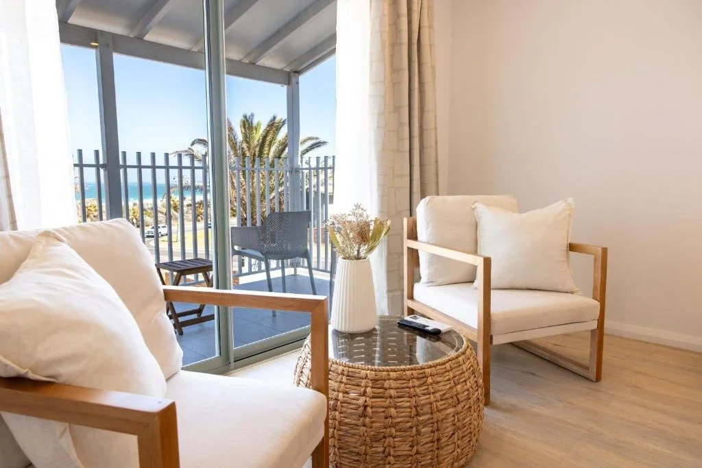 Living room with two white cushioned armchairs with wooden frames, a round wicker table with a vase of flowers and a remote, a sliding glass door opening to a balcony with outdoor chairs, a view of palm trees, a parking lot, and the ocean in the back