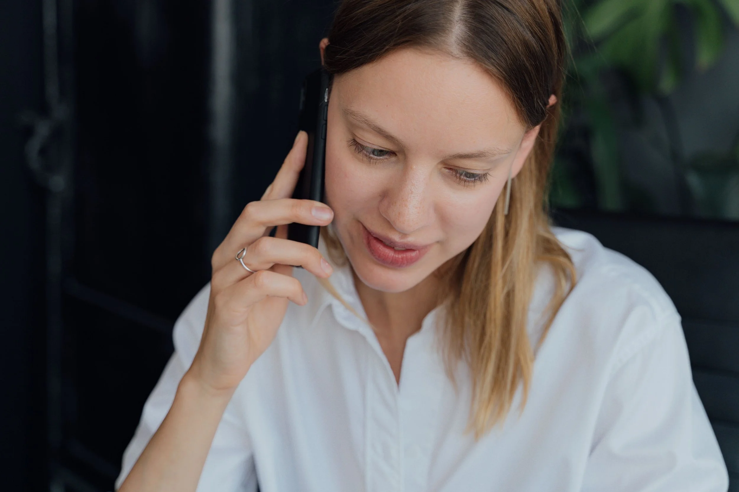 A young woman with red hair, wearing a white shirt, talking on a mobile phone. She has a silver ring on her finger and an earring. She appears to be indoors with dark background and greenery outside the window.