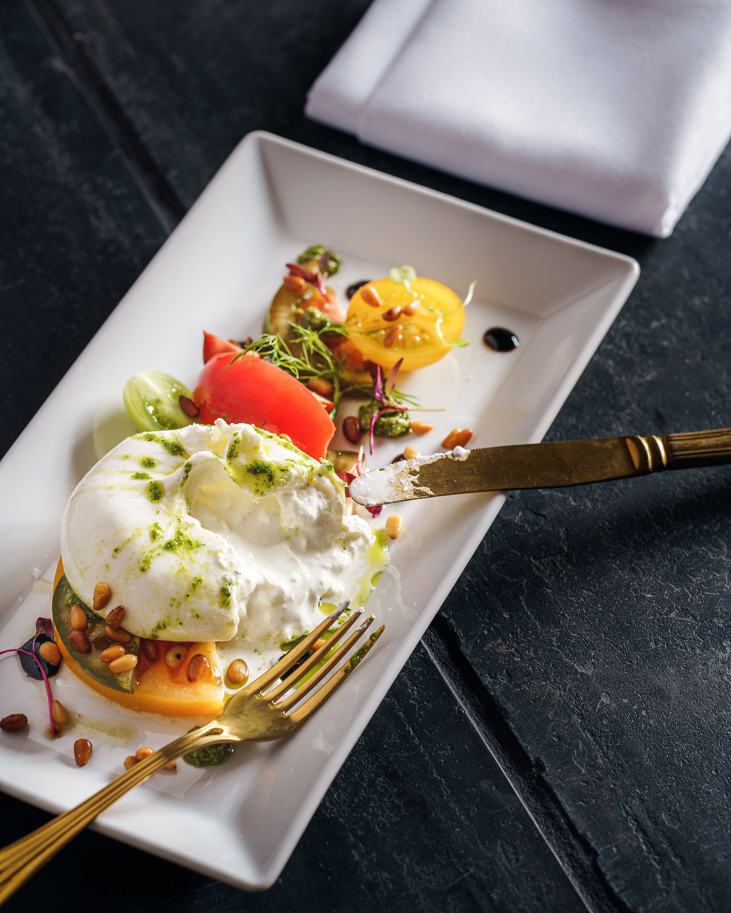 Close-up of a dessert plate with whipped cream, mango slices, and garnishes, on a dark textured surface, with a gold fork placed on the plate.