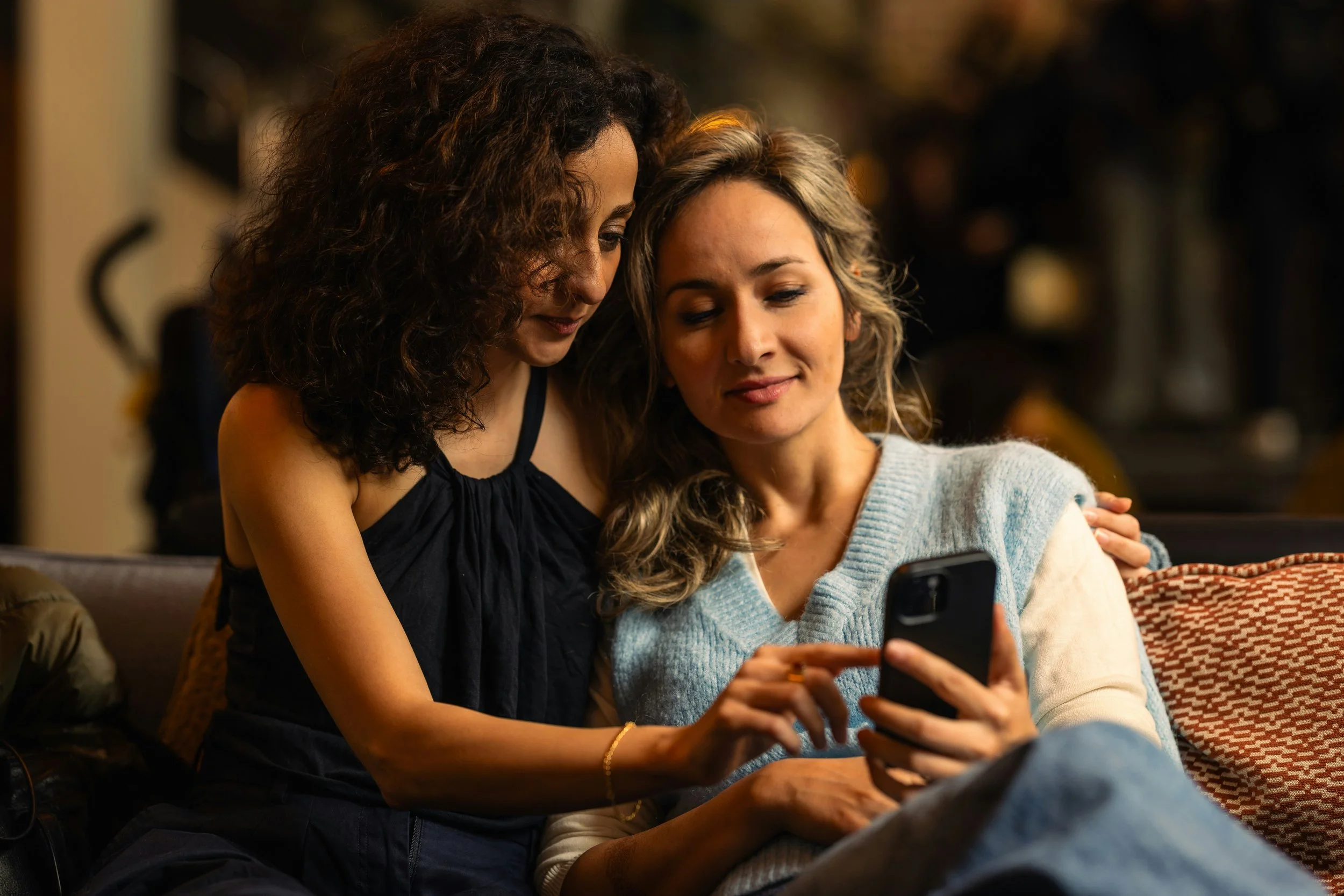 Two women sitting close together, one with curly dark hair wearing a black top, and one with wavy blonde hair wearing a light blue knit sweater, looking at a smartphone.