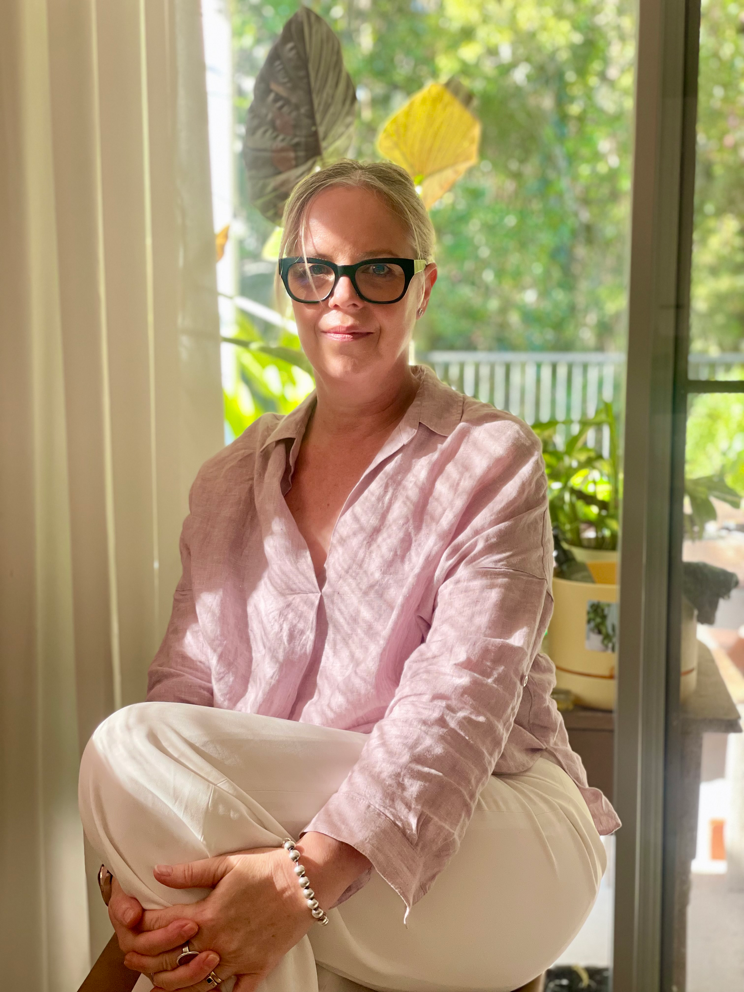 Woman with blonde hair wearing large black glasses and a light pink shirt, sitting indoors near a window with lush green foliage outside, holding her arms crossed with a silver bracelet on her wrist.