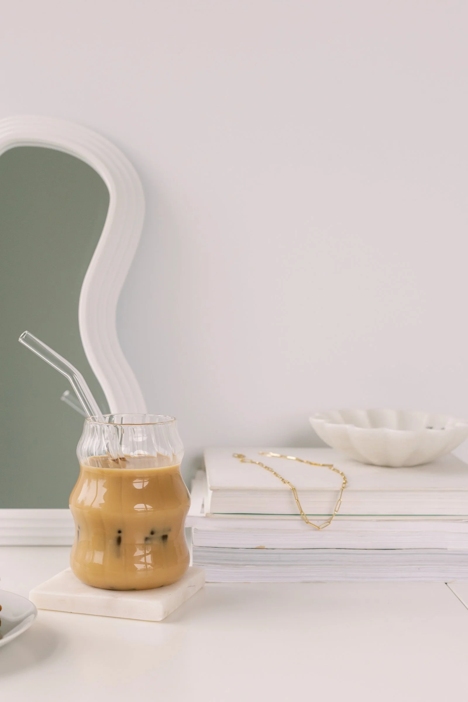 A pearl-beige ceramic vase with bubble-like design holding iced coffee with a straw, placed on a white marble coaster on a white surface. Behind it, a stack of porcelain whites and a gold chain necklace resting on a closed white book, with a white bowl in the background.