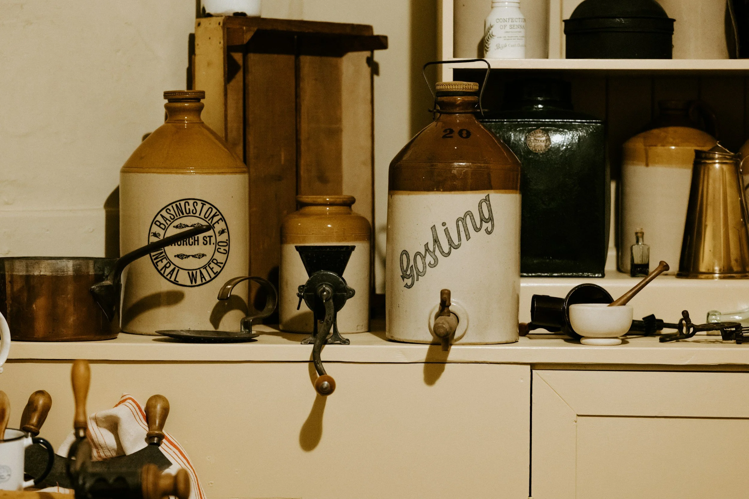 Vintage kitchen containers labeled 'basingstoke', 'ghosting', and 'confection of senna' on a cream-colored cabinet with various kitchen items.