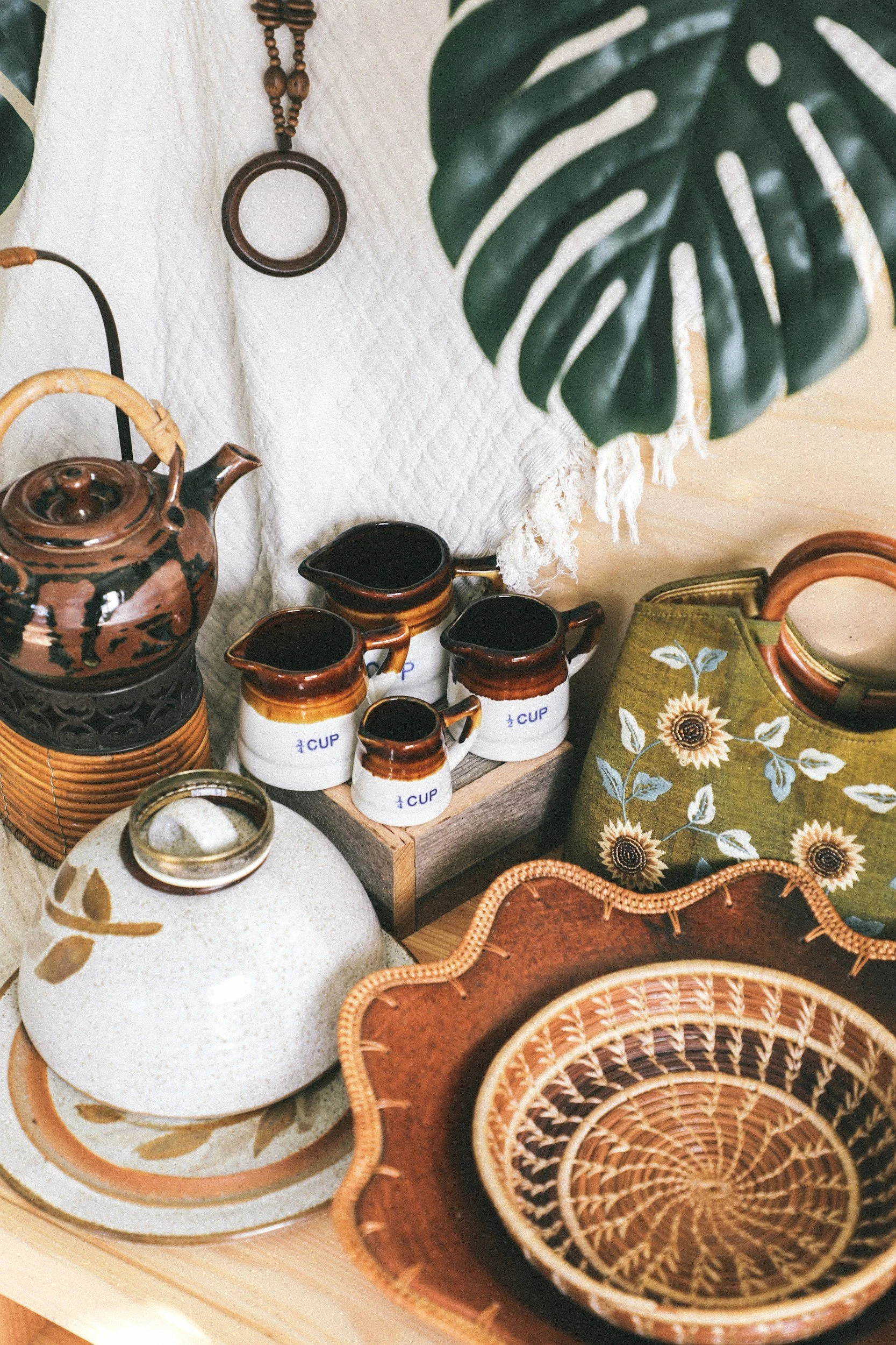 Decorative kitchen items including a ceramic teapot, small measuring cups, a woven tray, a green bag with sunflower pattern, and a glass jar, with a large green monstera leaf overhead.