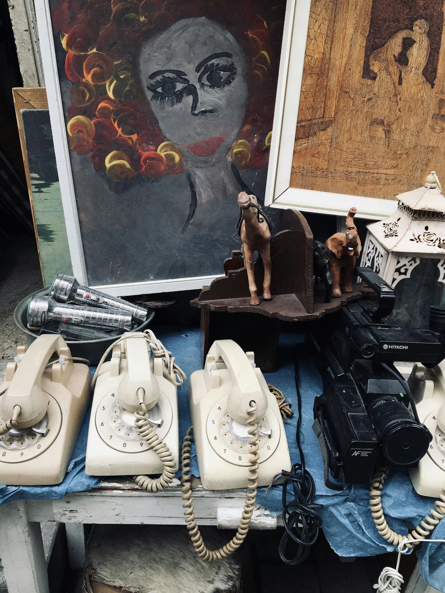 A collection of vintage rotary phones, a video camera, and various decorative items including framed artwork depicting a woman with colorful hair, wooden sculpture figures, and a white lantern.