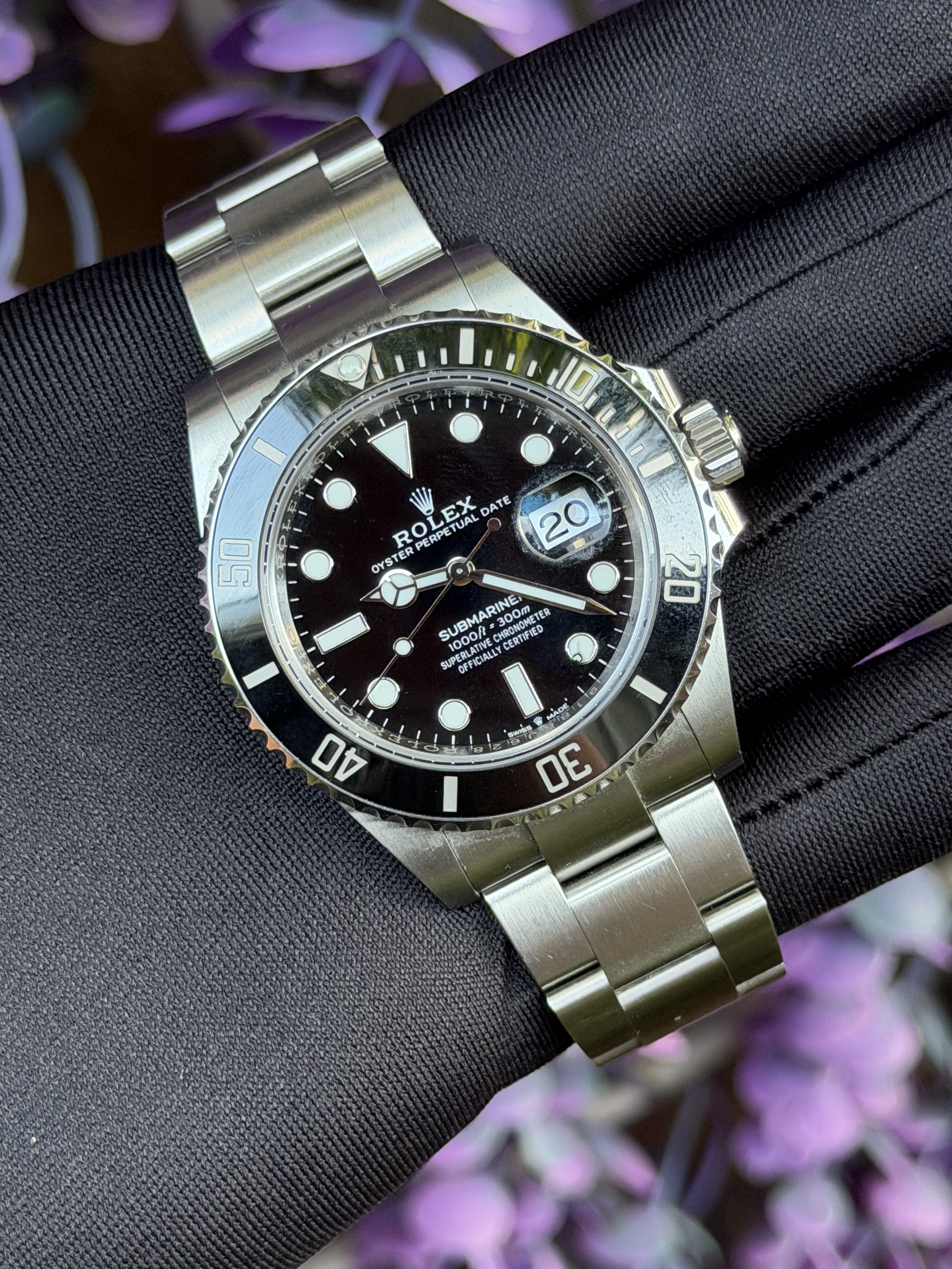 Silver Rolex Submariner watch with black dial and bezel, being held in a gloved hand with blurred pink and purple flowers in the background.