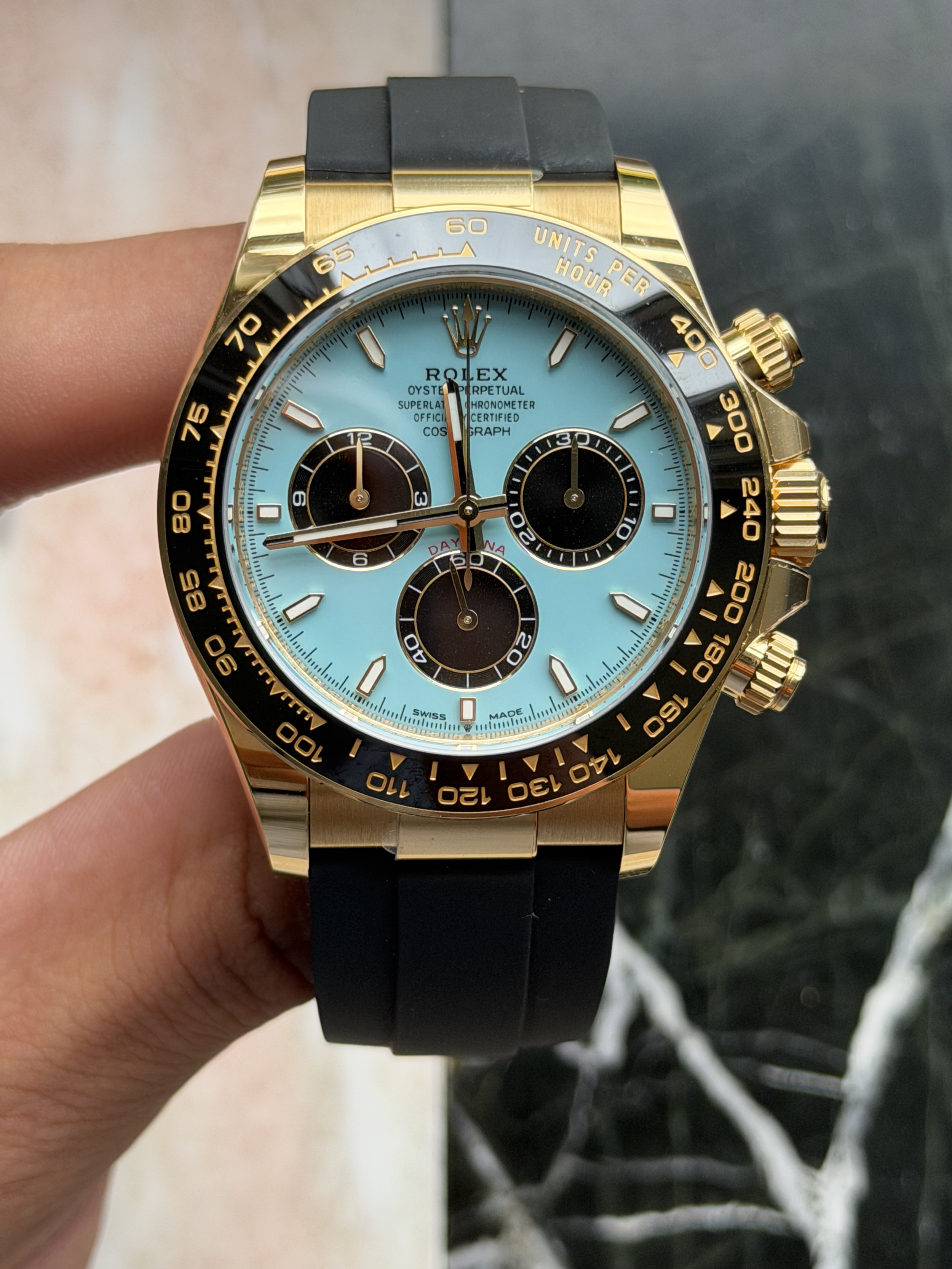 A gold and black Rolex Cosmograph Daytona watch with a light blue dial, black subdials, and a black rubber strap.