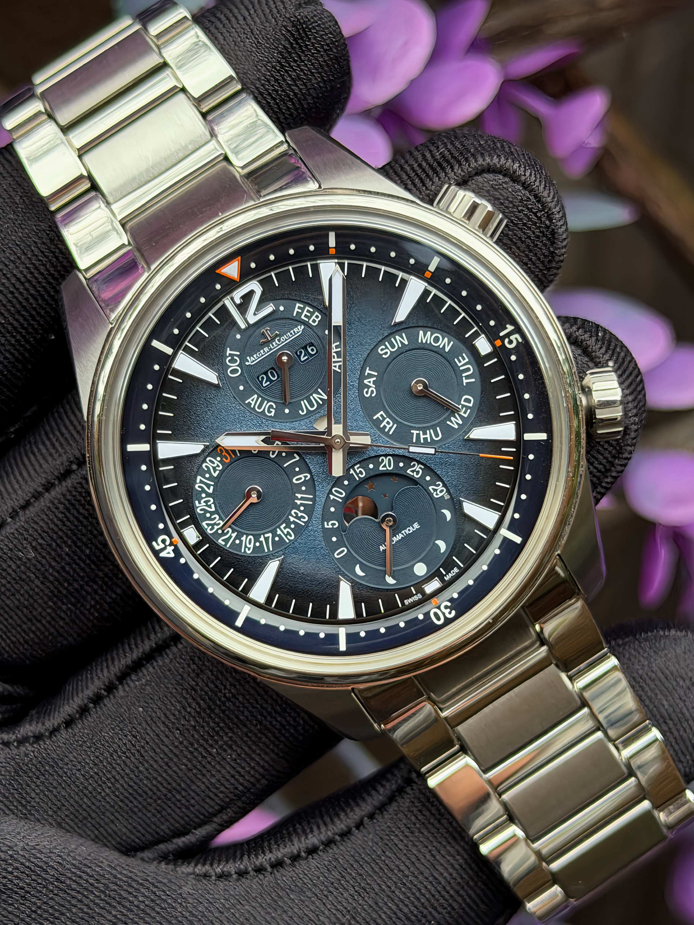 Close-up of a luxury wristwatch with a stainless steel band, multiple dials indicating date, days, months, and moon phase, held by a gloved hand with purple flowers in the background.