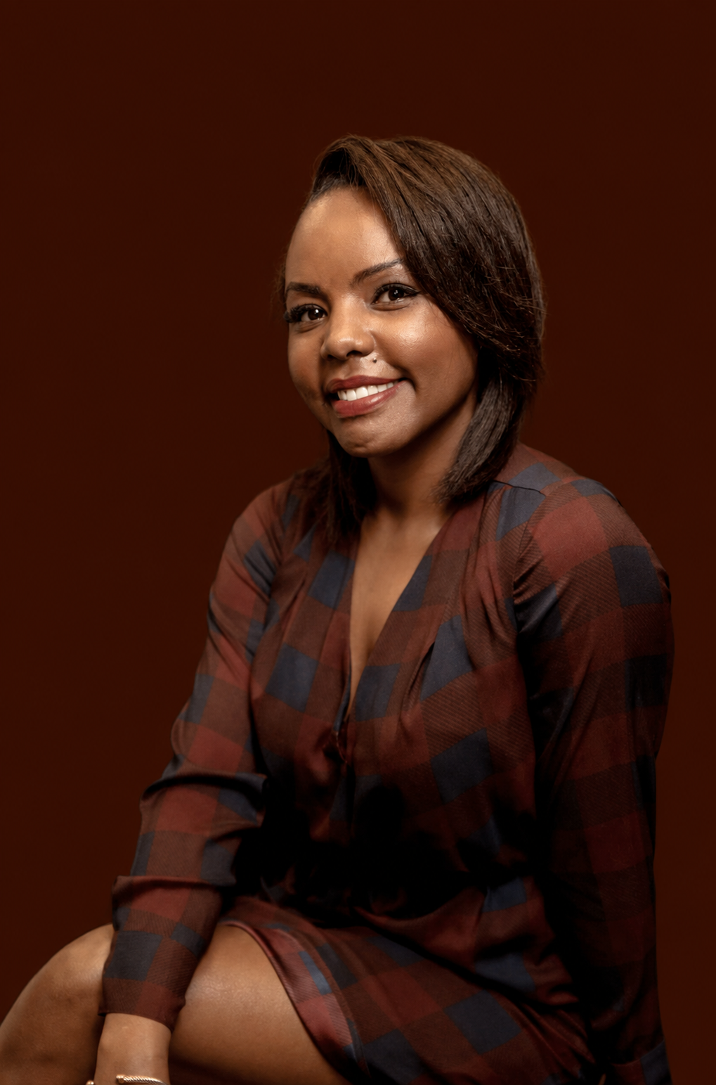 A woman with shoulder-length dark hair, wearing a brown and black checkered dress, sitting against a brown background, smiling at the camera.