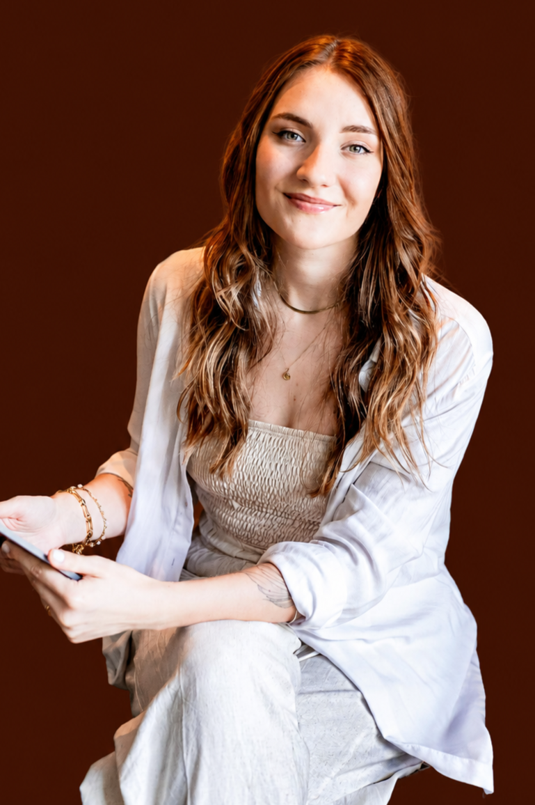 Young woman with long wavy hair sitting and smiling, holding a phone, against a brown background.