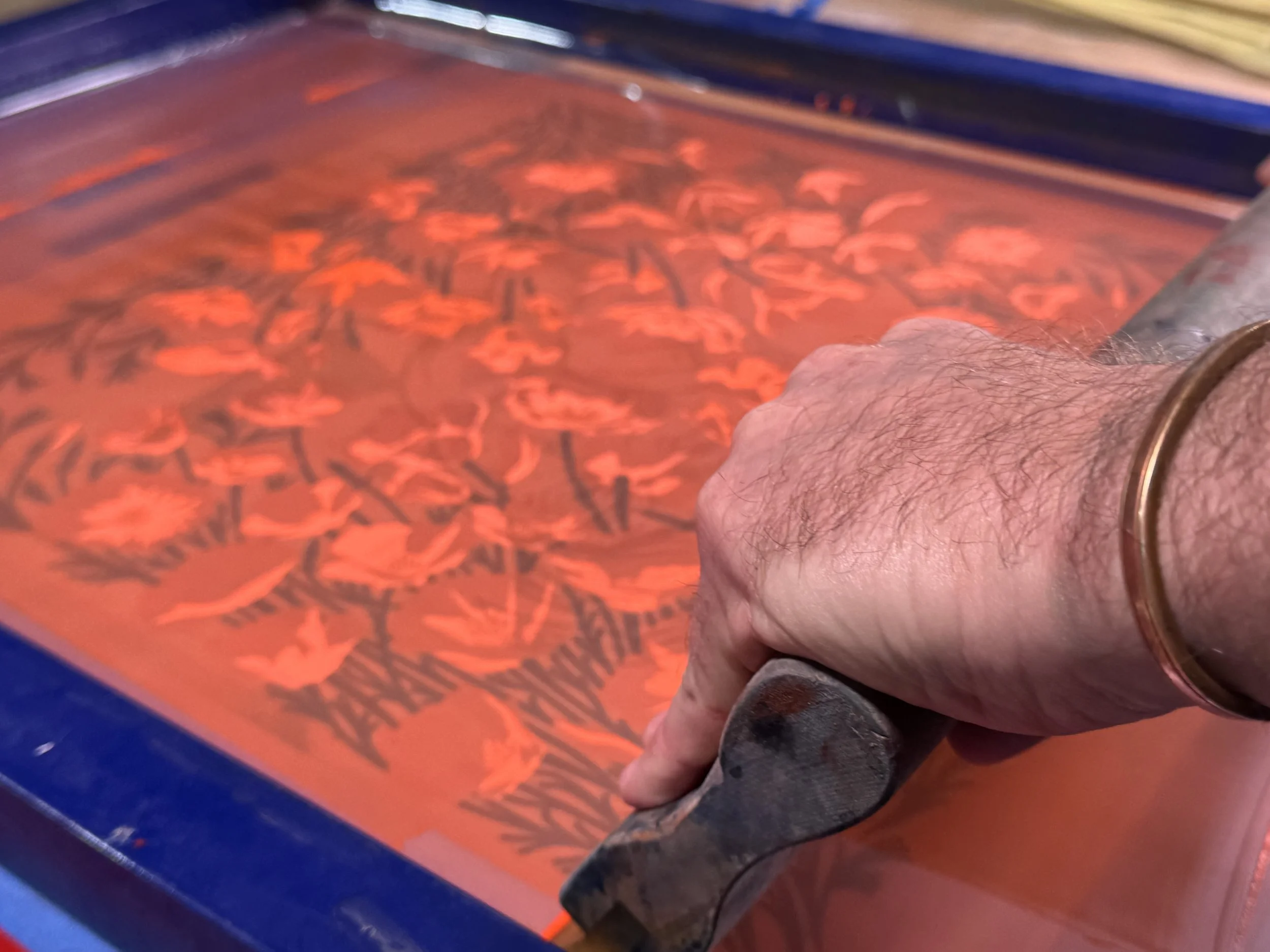 A person's hand using a squeegee to screenprint an orange and black design on a surface, likely a screen printing process.