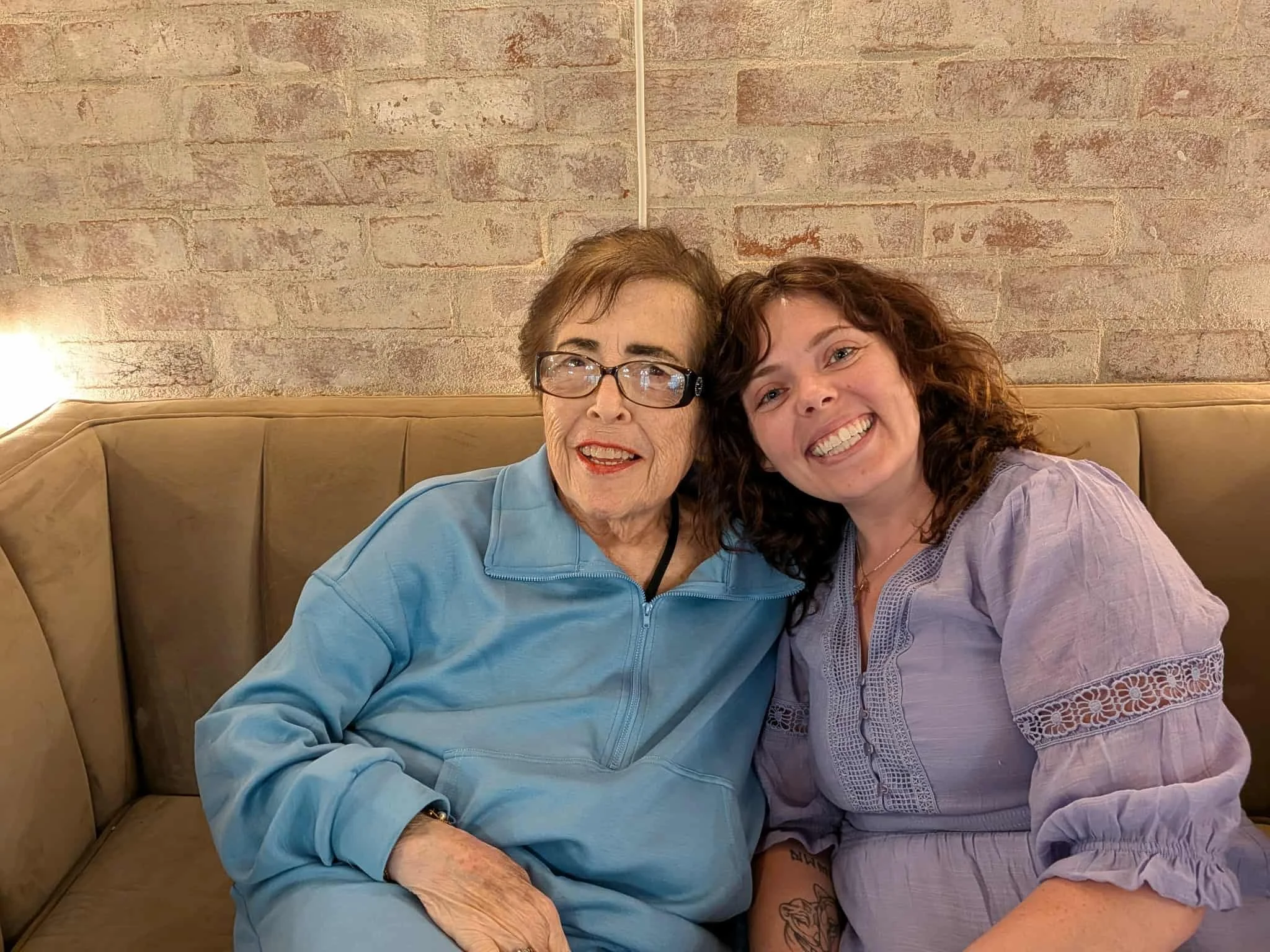 Two women sitting close together on a beige couch in front of a brick wall, smiling at the camera, one with glasses and the other with curly hair.