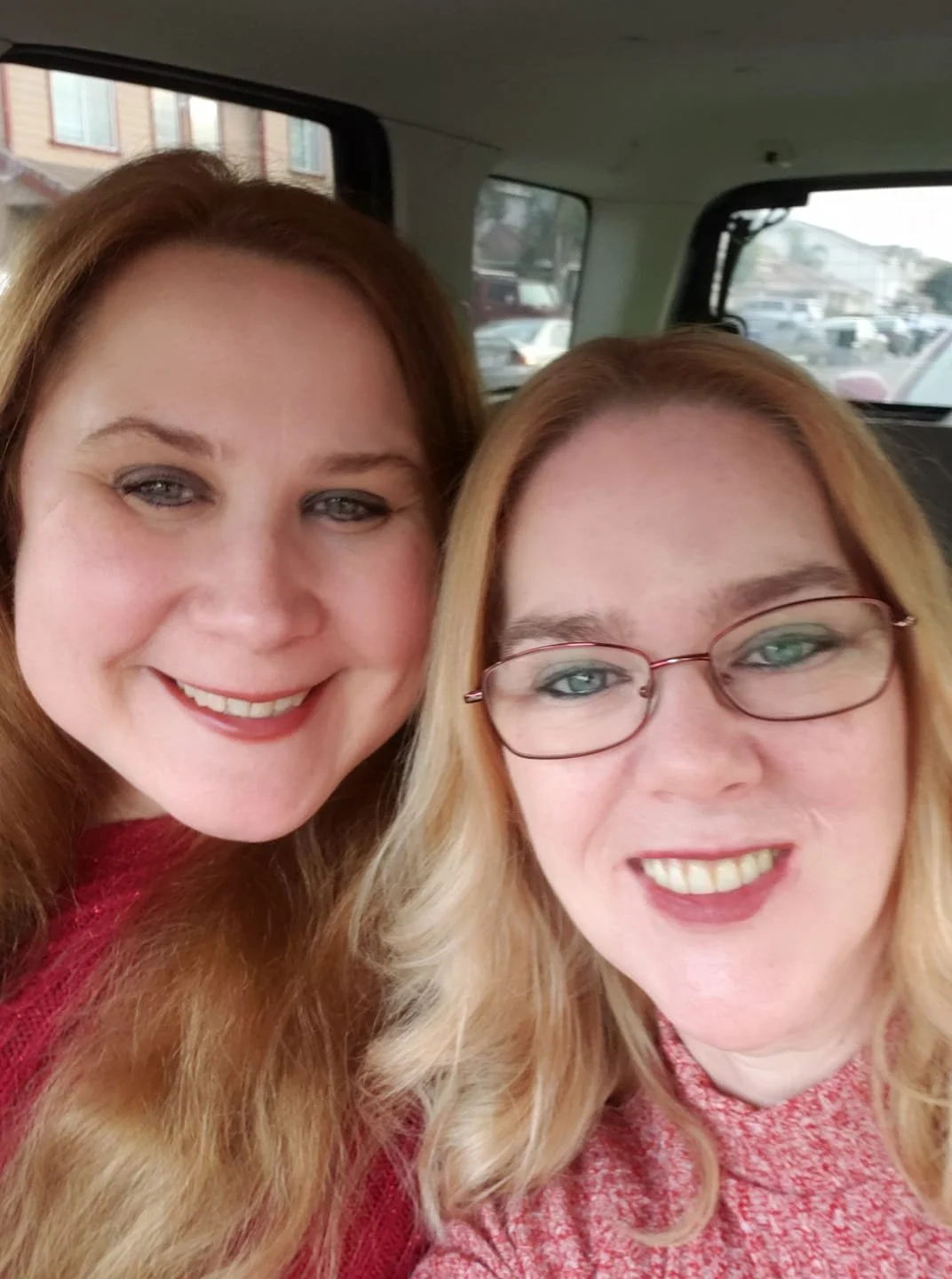 Two smiling women with long hair sitting in a vehicle, taking a selfie together.