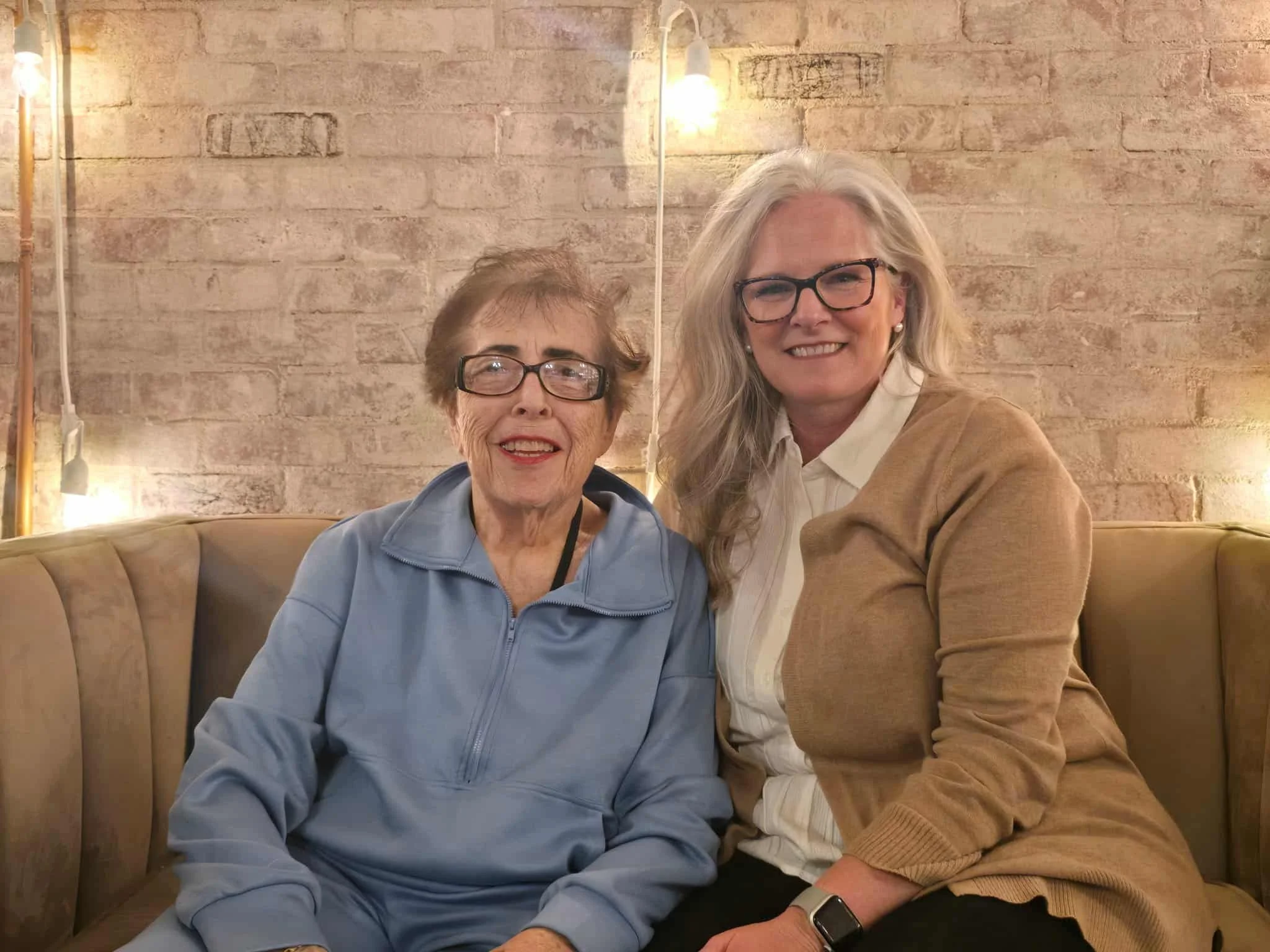 Two smiling women sitting on a beige sofa against a brick wall, wearing glasses, with warm lighting in the background.