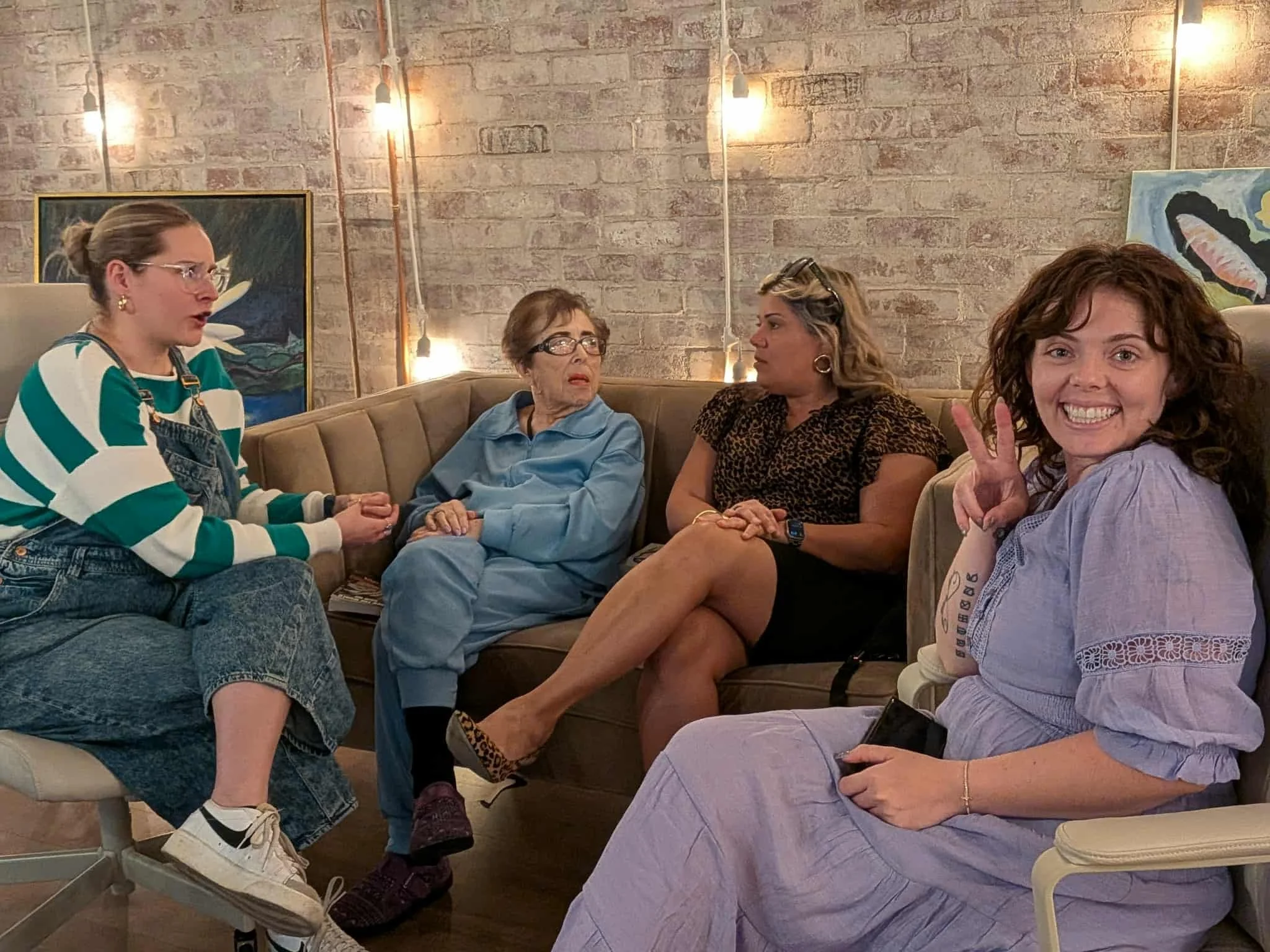 Five women sitting on a sofa in a cozy room with exposed brick walls and artwork, having a conversation. One woman is smiling and showing a peace sign.