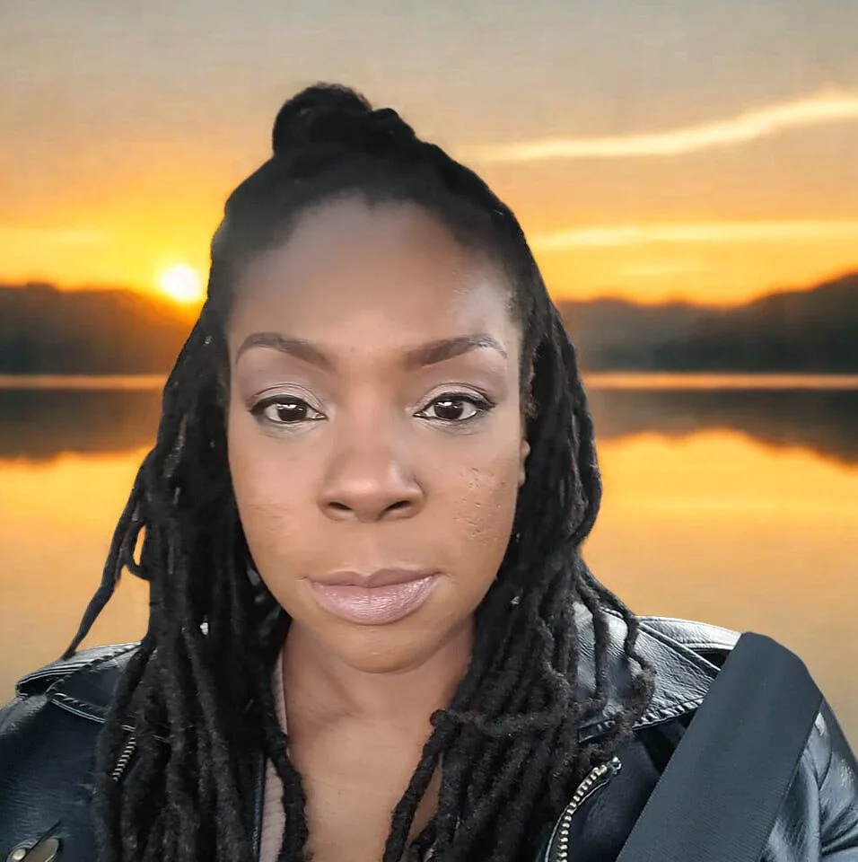 A woman with dreadlocks and a leather jacket taking a selfie with a sunset over a lake in the background.