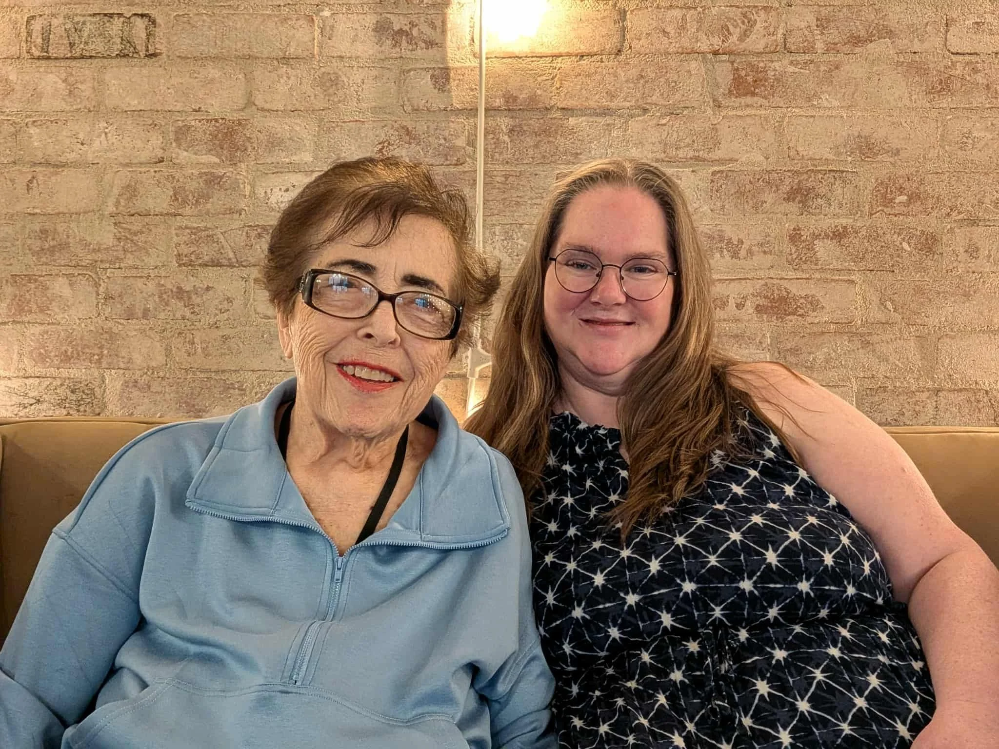 Two women smiling, sitting on a beige bench against a brick wall, with a lamp behind them. One woman is elderly with short brown hair, glasses, wearing a light blue zip-up top. The other woman is younger with long red hair, glasses, wearing a sleeveless black dress with a white star pattern.