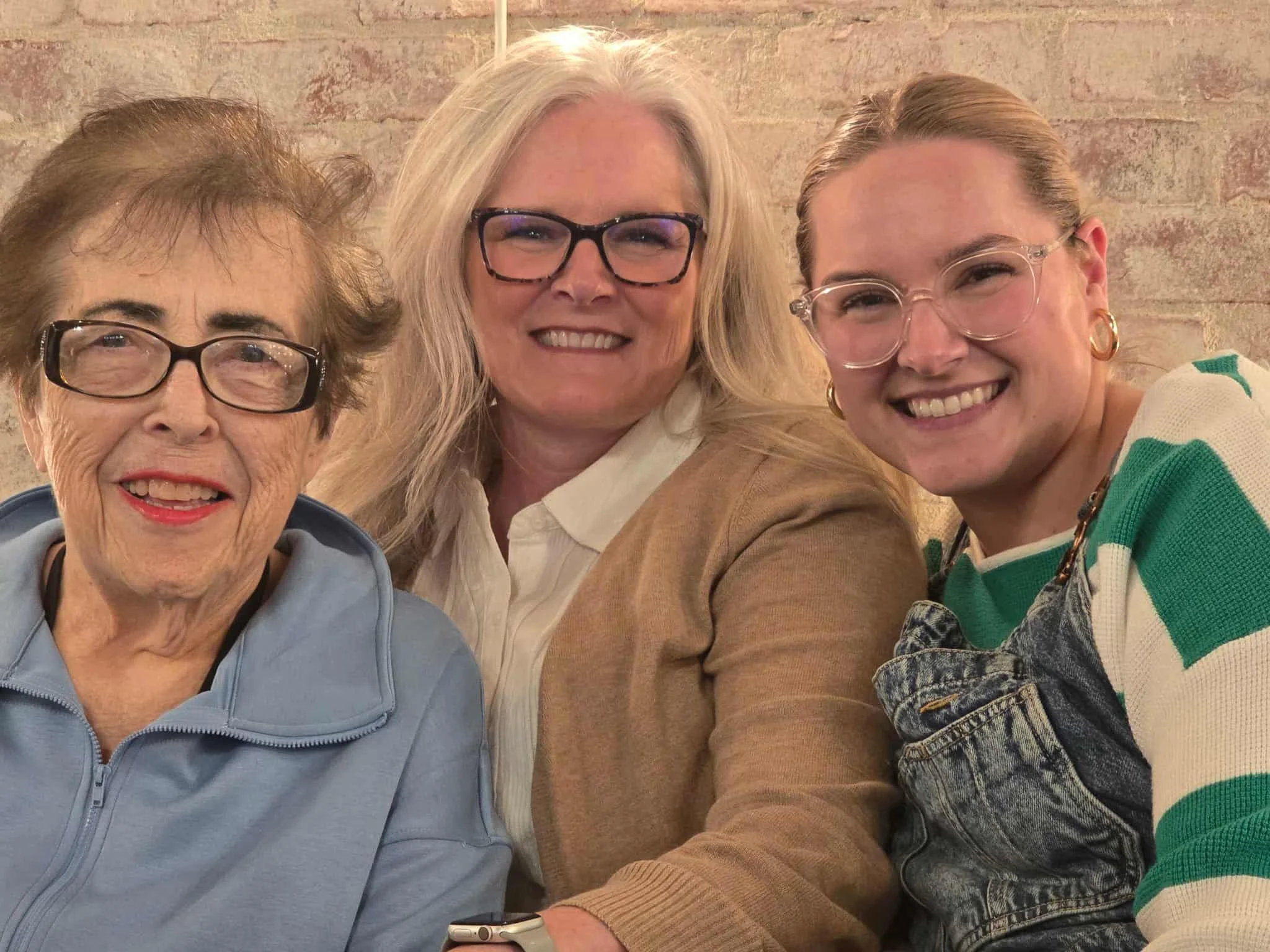 Three women smiling for a selfie in front of a brick wall. The woman on the left has short brown hair, glasses, and is wearing a light blue zip-up jacket. The woman in the middle has long gray hair, glasses, and is wearing a white shirt with a tan cardigan. The woman on the right has pulled-back hair, glasses, gold hoop earrings, and is dressed in a striped green and white shirt with overalls.