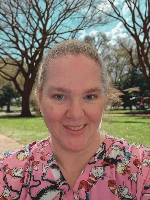 A woman with blonde hair smiling outdoors in front of leafless trees and a park with green grass.