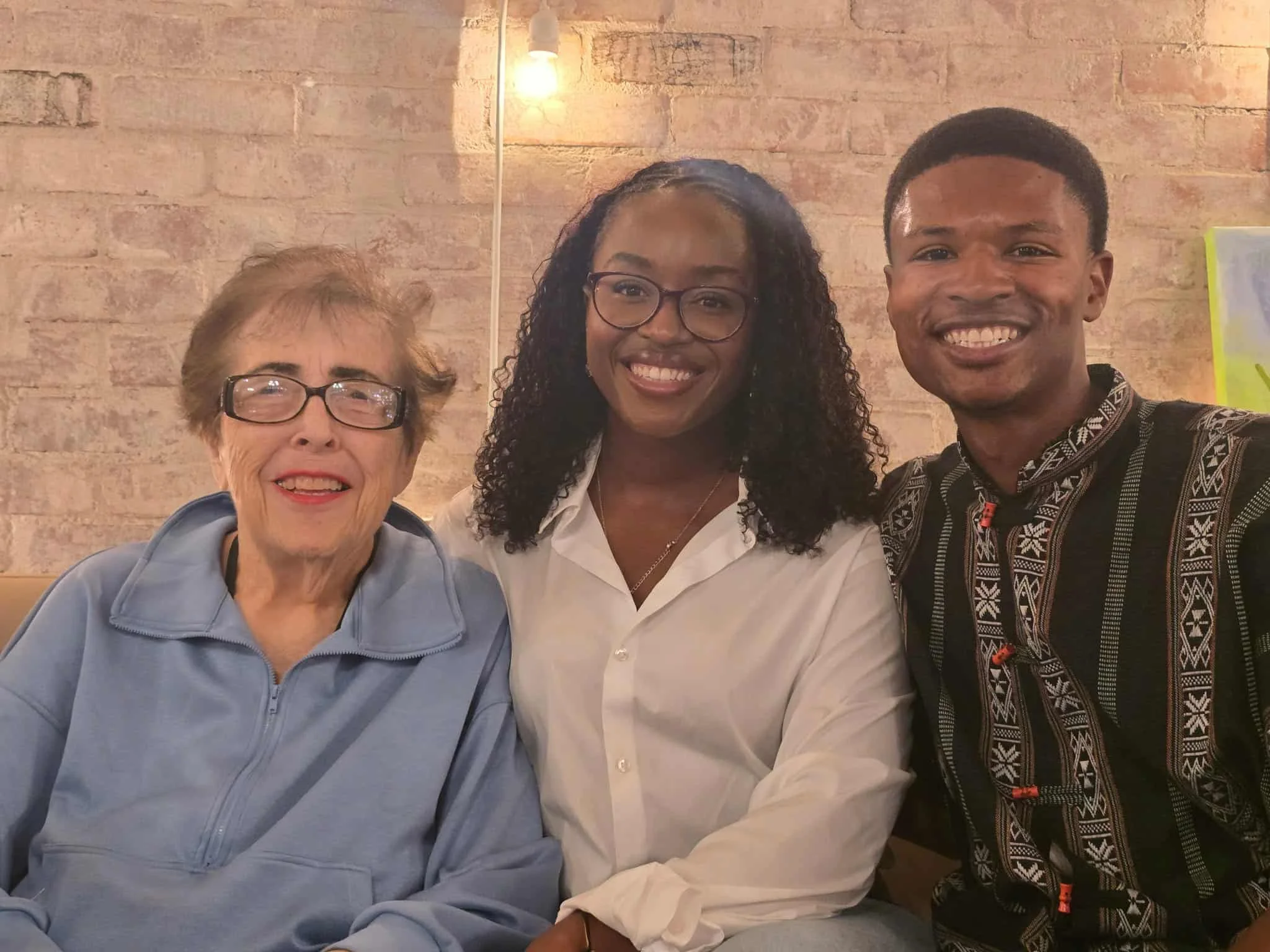 Three smiling women of different ages and ethnicities sitting together indoors, with a brick wall and a hanging light in the background.