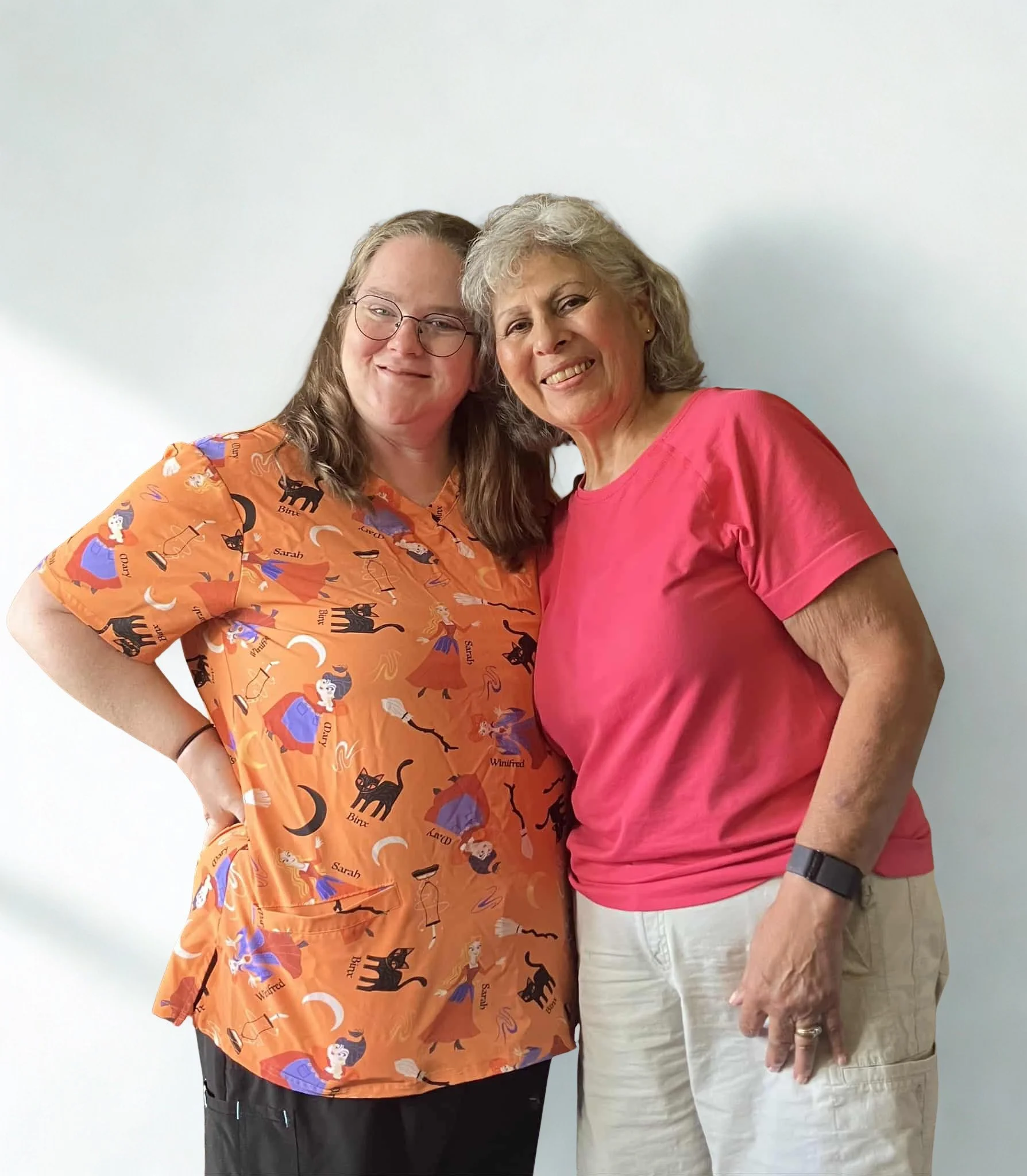 Two women smiling and standing close together against a plain white wall.
