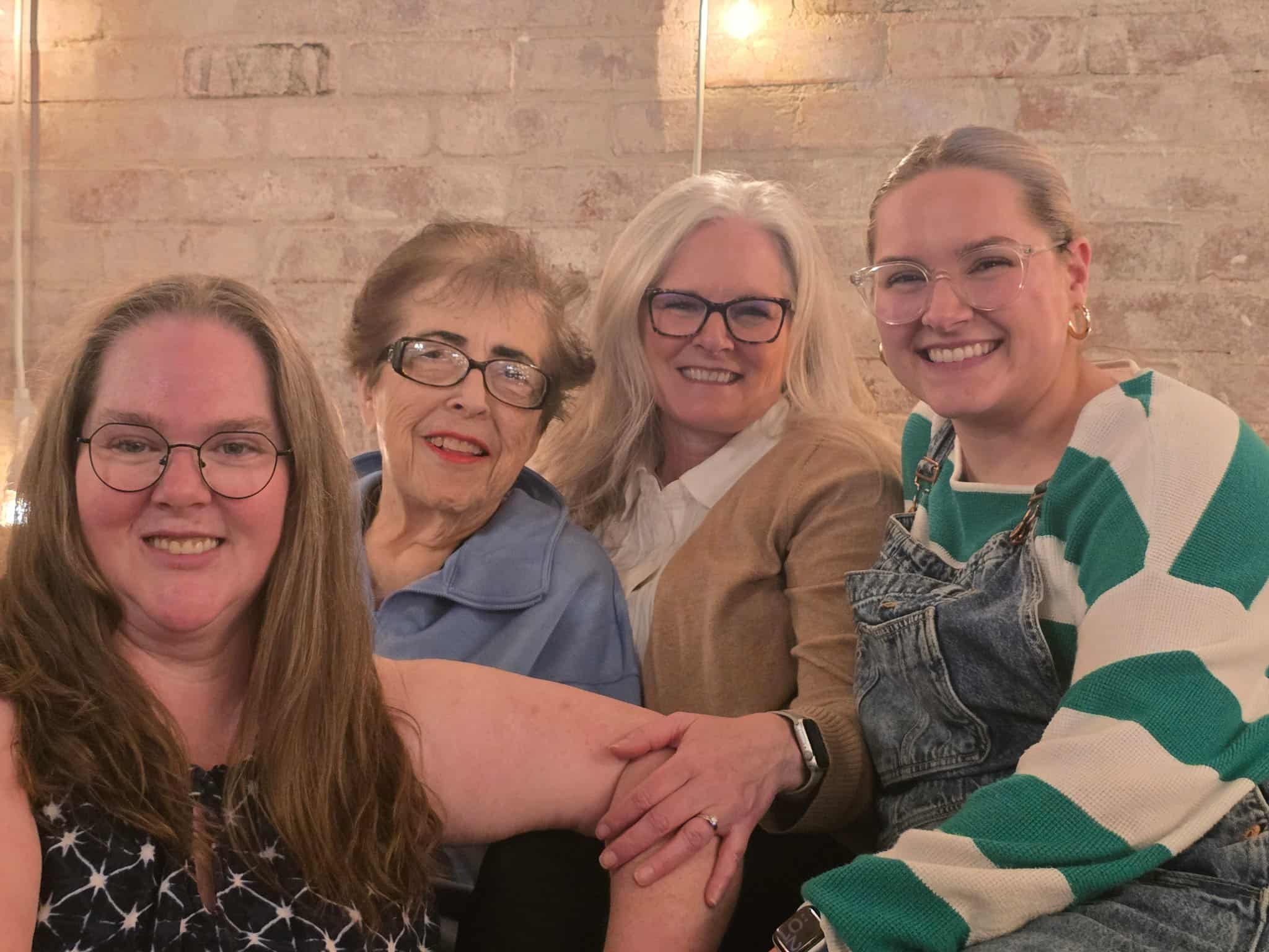 Group of four women with diverse hair colors and styles, smiling and holding hands, in a warmly lit indoor setting with a brick wall background.
