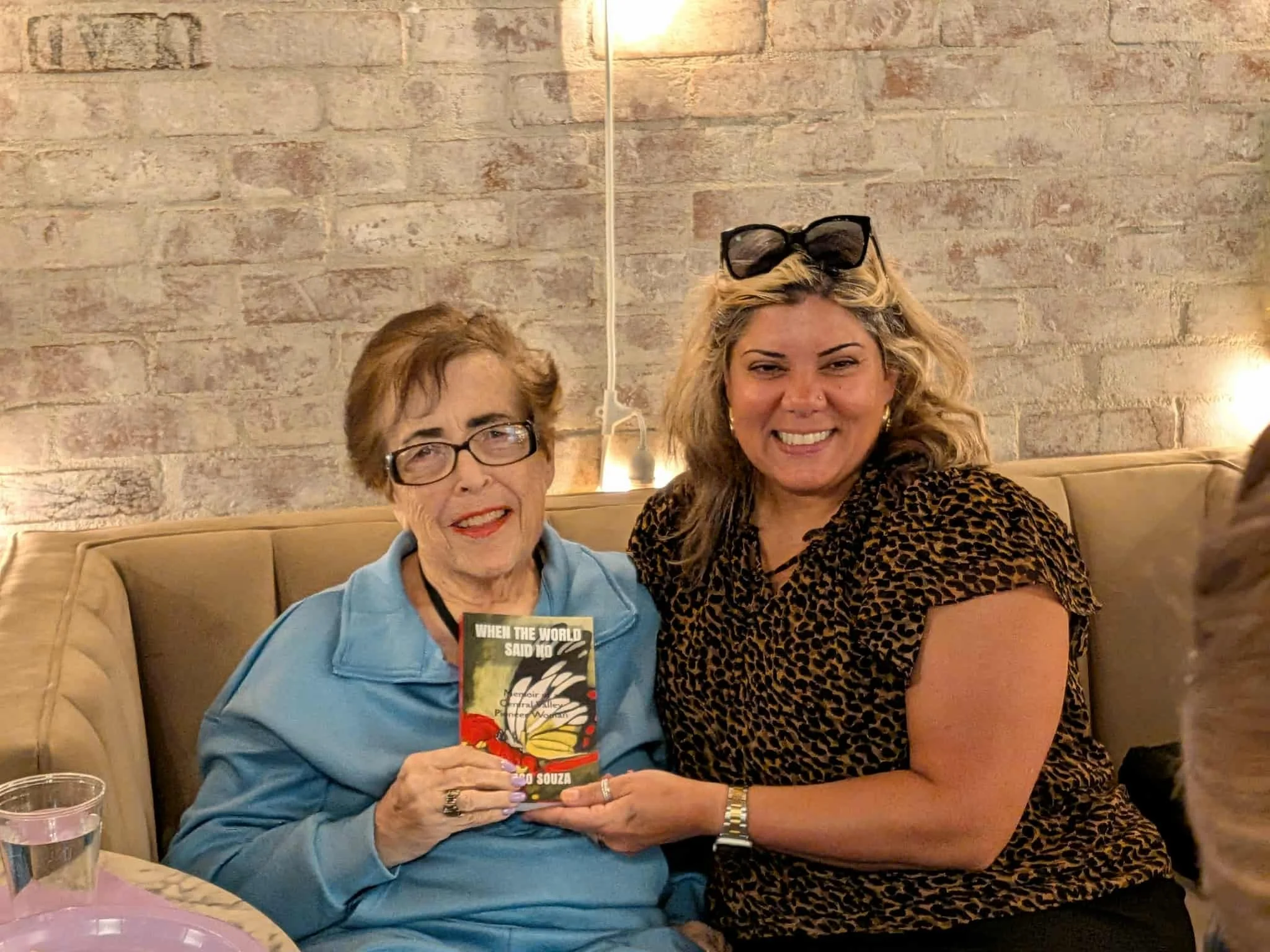 Two women sitting on a beige booth in a restaurant, smiling at the camera, one holding a book titled 'When the World Said No'. The woman on the left has short brown hair, glasses, and is wearing a blue jacket, while the woman on the right has long blonde hair, sunglasses on her head, and is wearing a leopard print top.