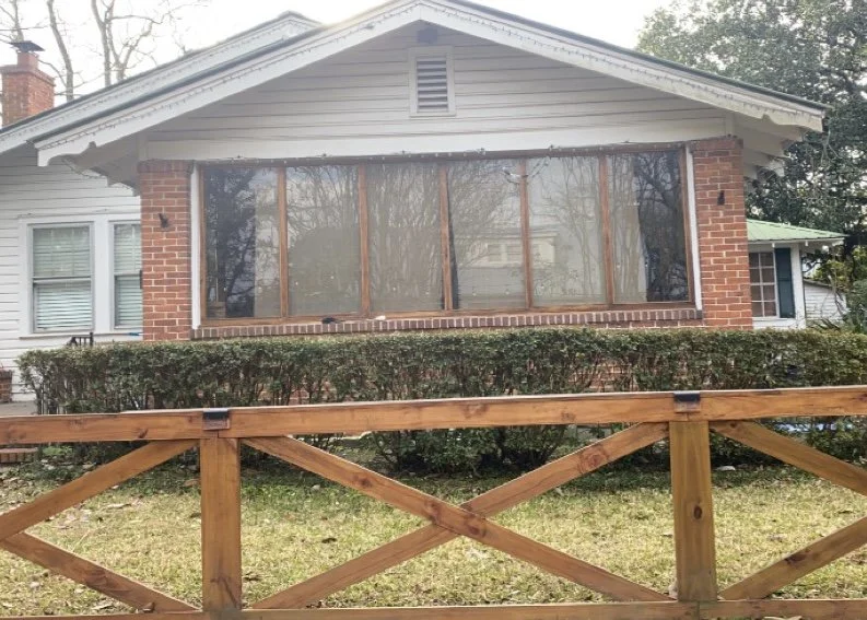A house with a large front window and a small hedge in front, with a wooden fence in the foreground.