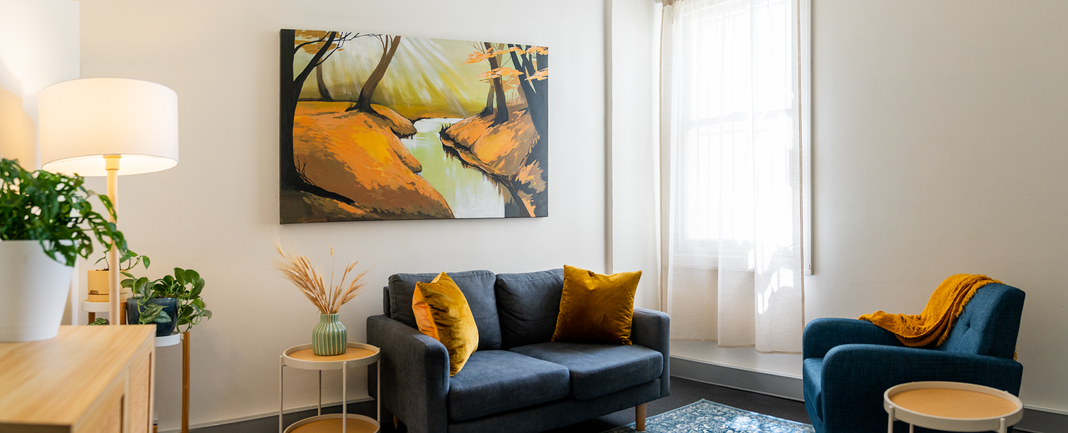 Consulting room with a grey sofa, blue armchair, yellow cushions and natural light.