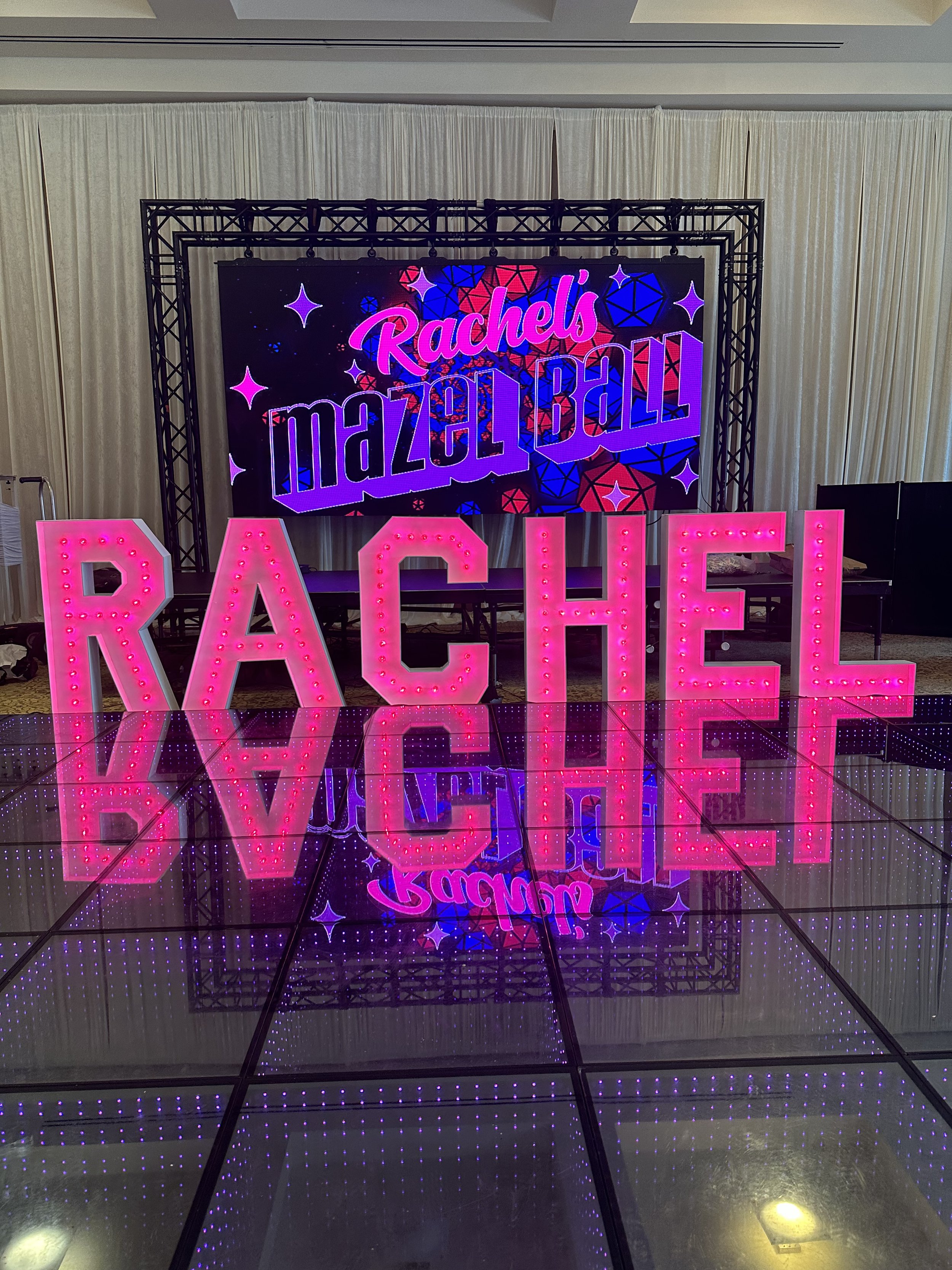 Bright pink illuminated letters spell "RACHEL" in front of a screen that reads "Rachel's Maze Ball" with colorful geometric patterns and stars.