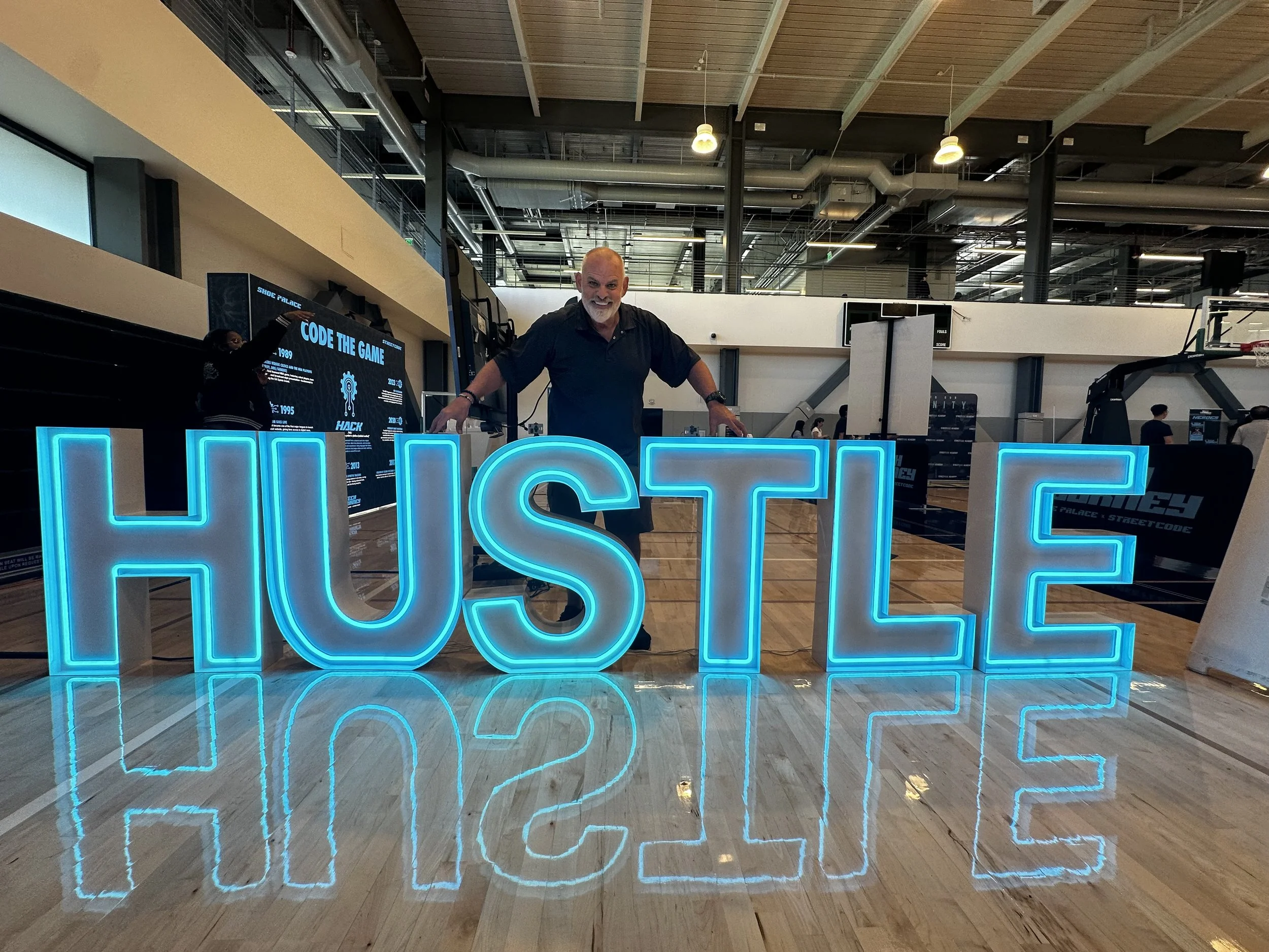 A man standing behind illuminated blue sign spelling 'HUSTLE' with a gym or sports hall in the background.