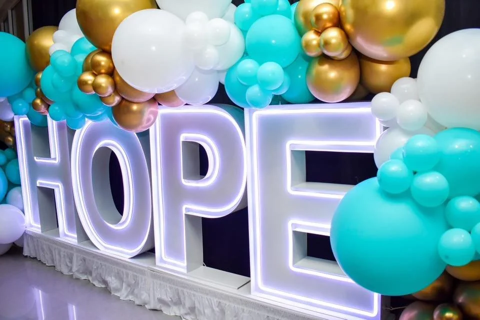 Large illuminated letters spelling 'HOPE' with colorful balloons in white, teal, gold, and pink surrounding them.