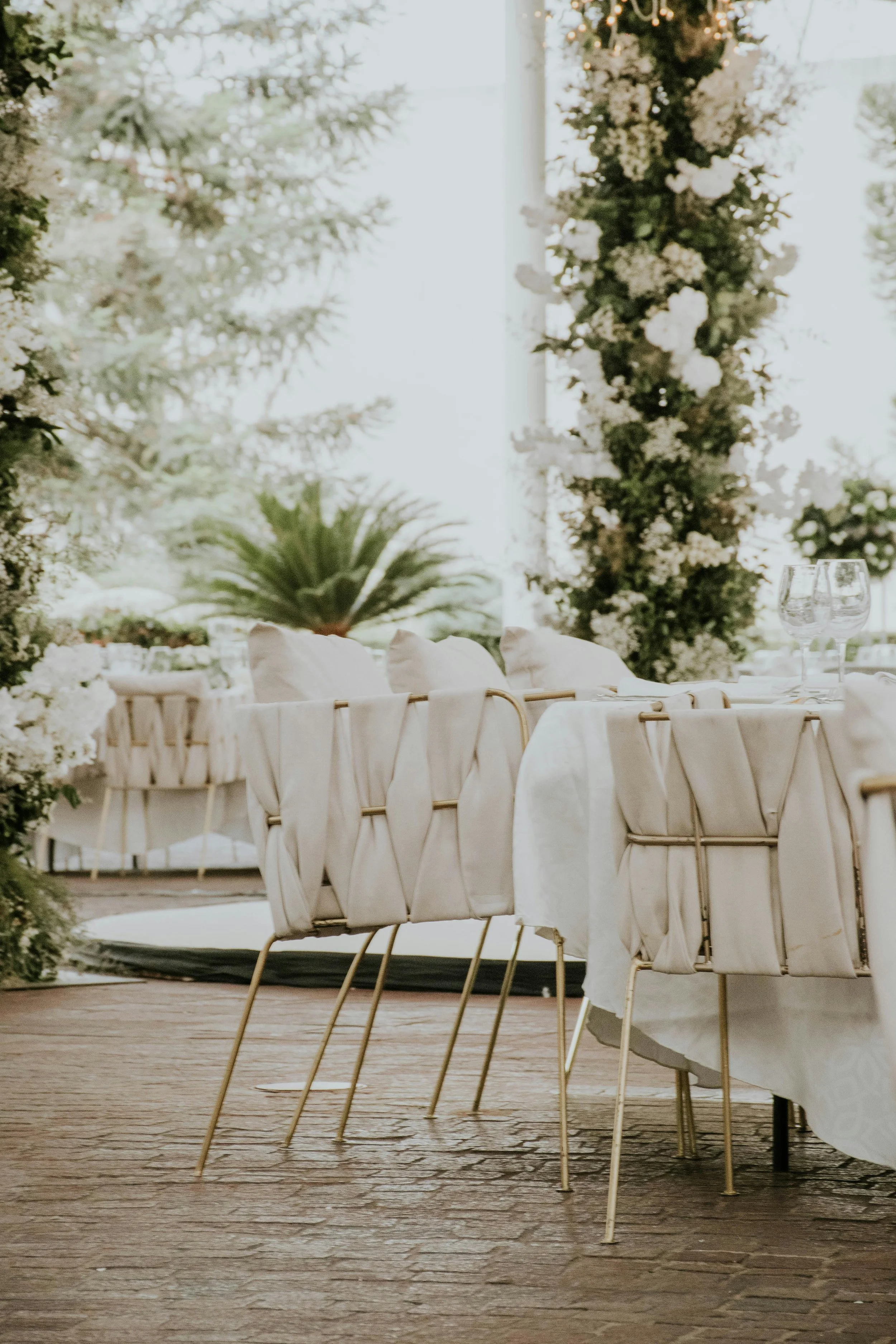 Elegant outdoor dining setting with white chairs, tablecloths, and floral decor under a clear tent with greenery in the background.
