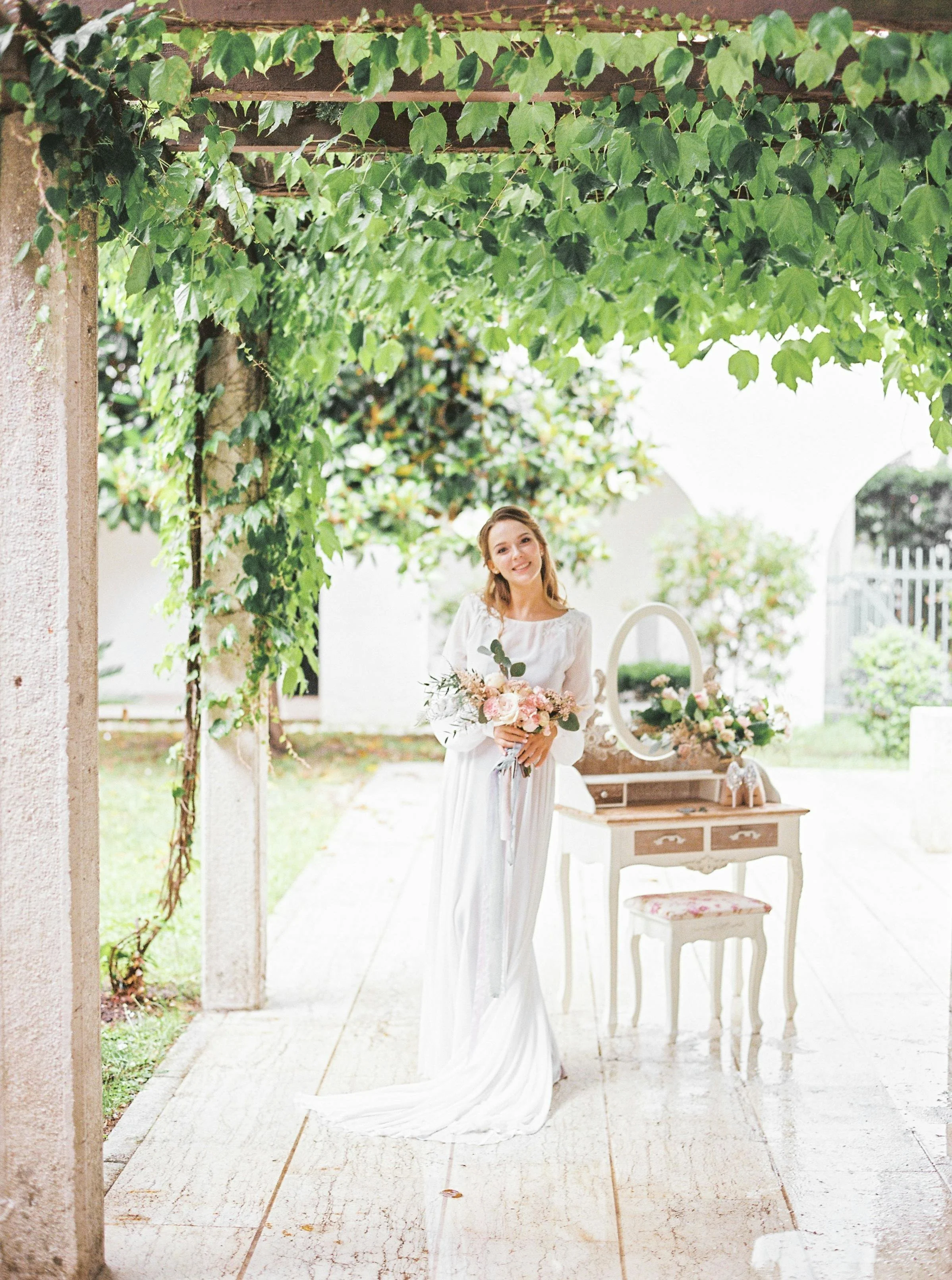 A woman in a white dress holding a bouquet of flowers standing under a leafy green arbor in a bright outdoor setting with a vintage vanity table and mirror in the background.