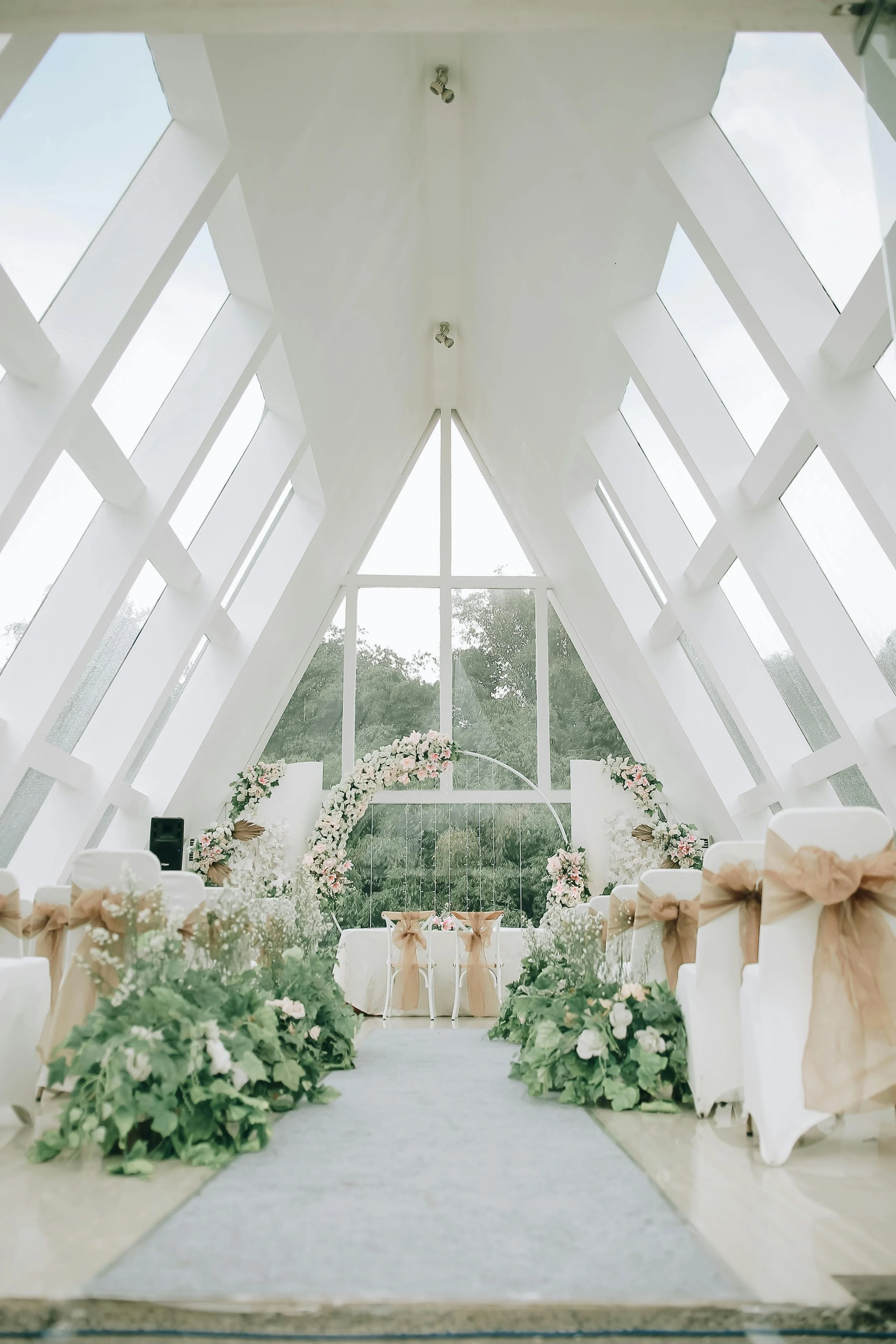A beautifully decorated indoor wedding ceremony space with white chairs adorned with beige ribbons, green foliage, and floral arrangements leading to an altar with a floral arch and a table in front of large glass windows.