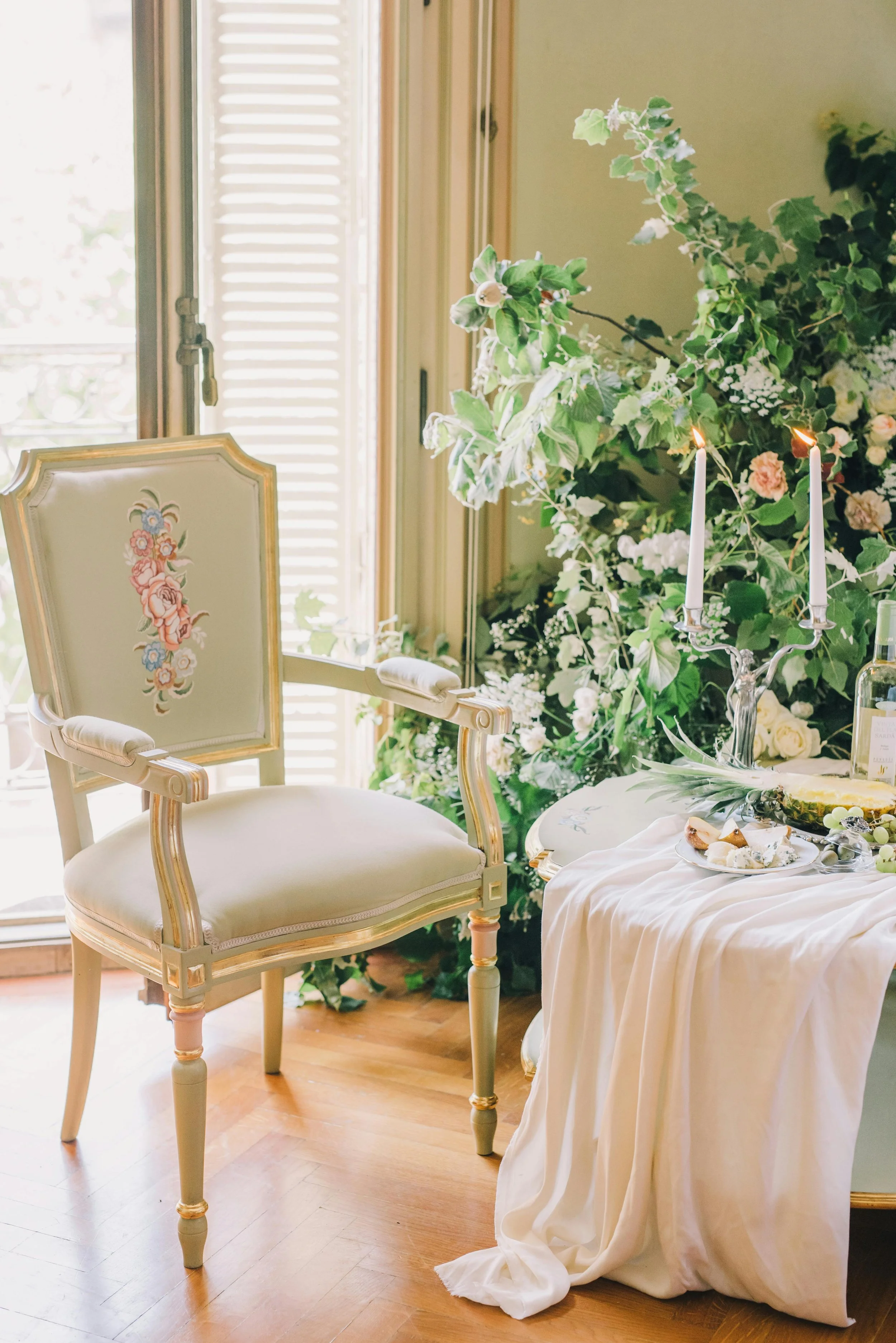 Elegant room with cream-colored chair with floral embroidery, next to a window with wooden shutters, and a table with a white cloth, lit candles, and floral decorations.