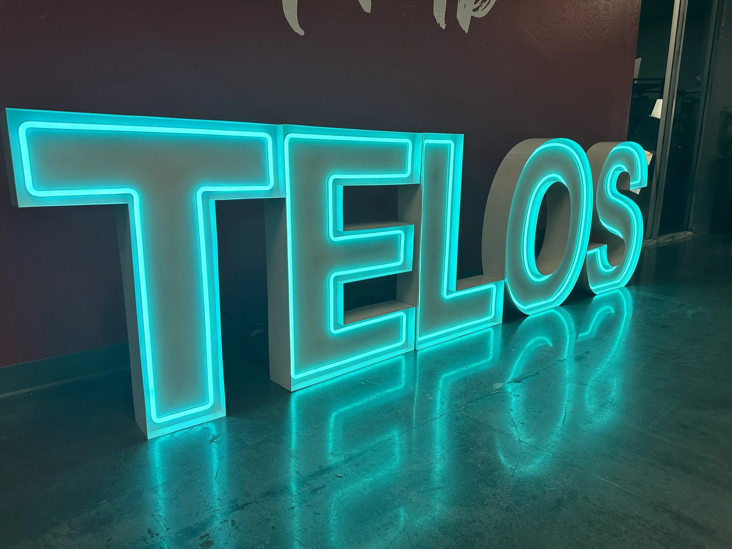 Bright blue neon sign spelling 'TELOS' on a dark wall, with a reflection on the floor.