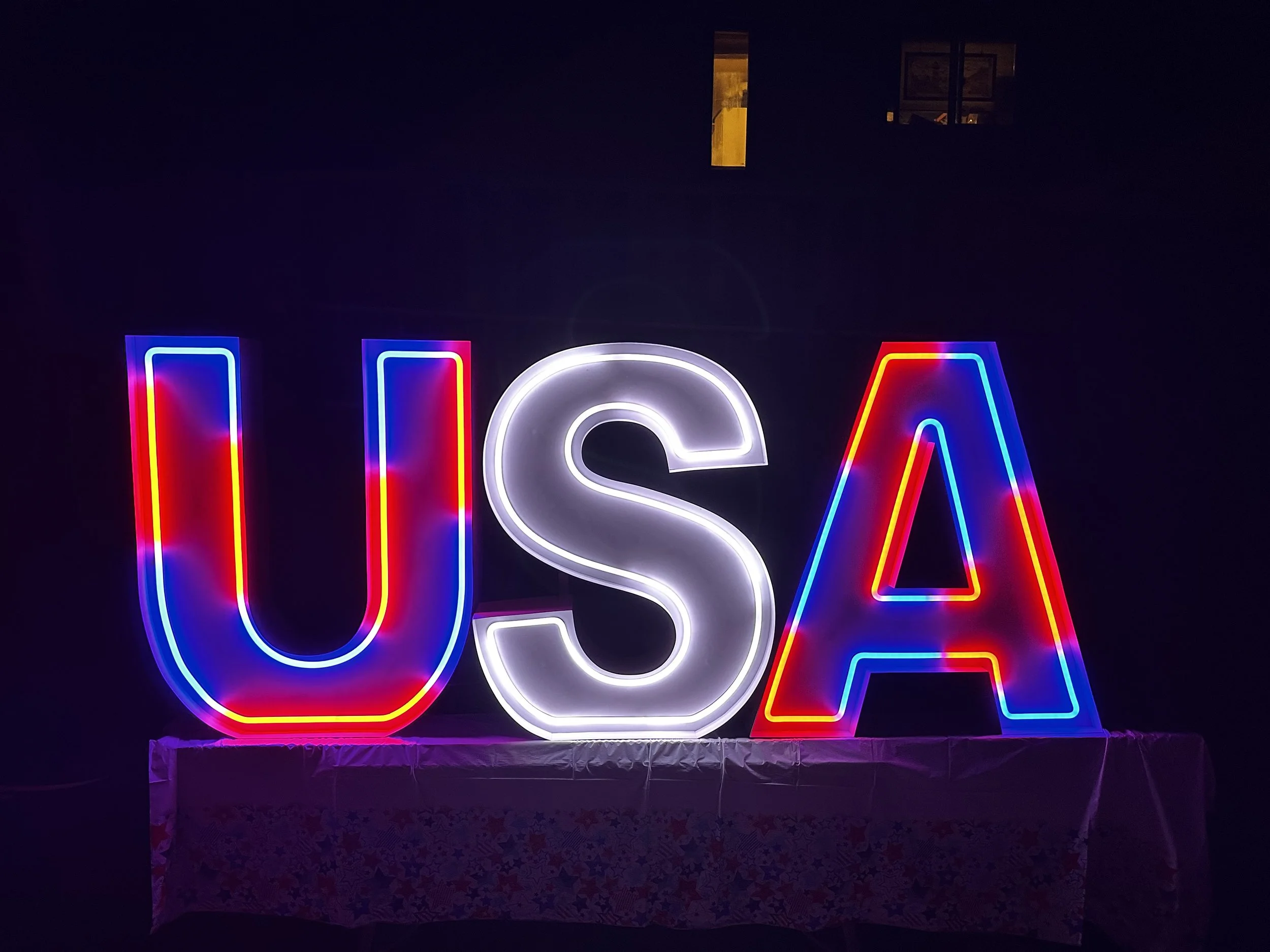 Neon sign spelling out "USA" with the letters illuminated in red, white, and blue, on a dark background.