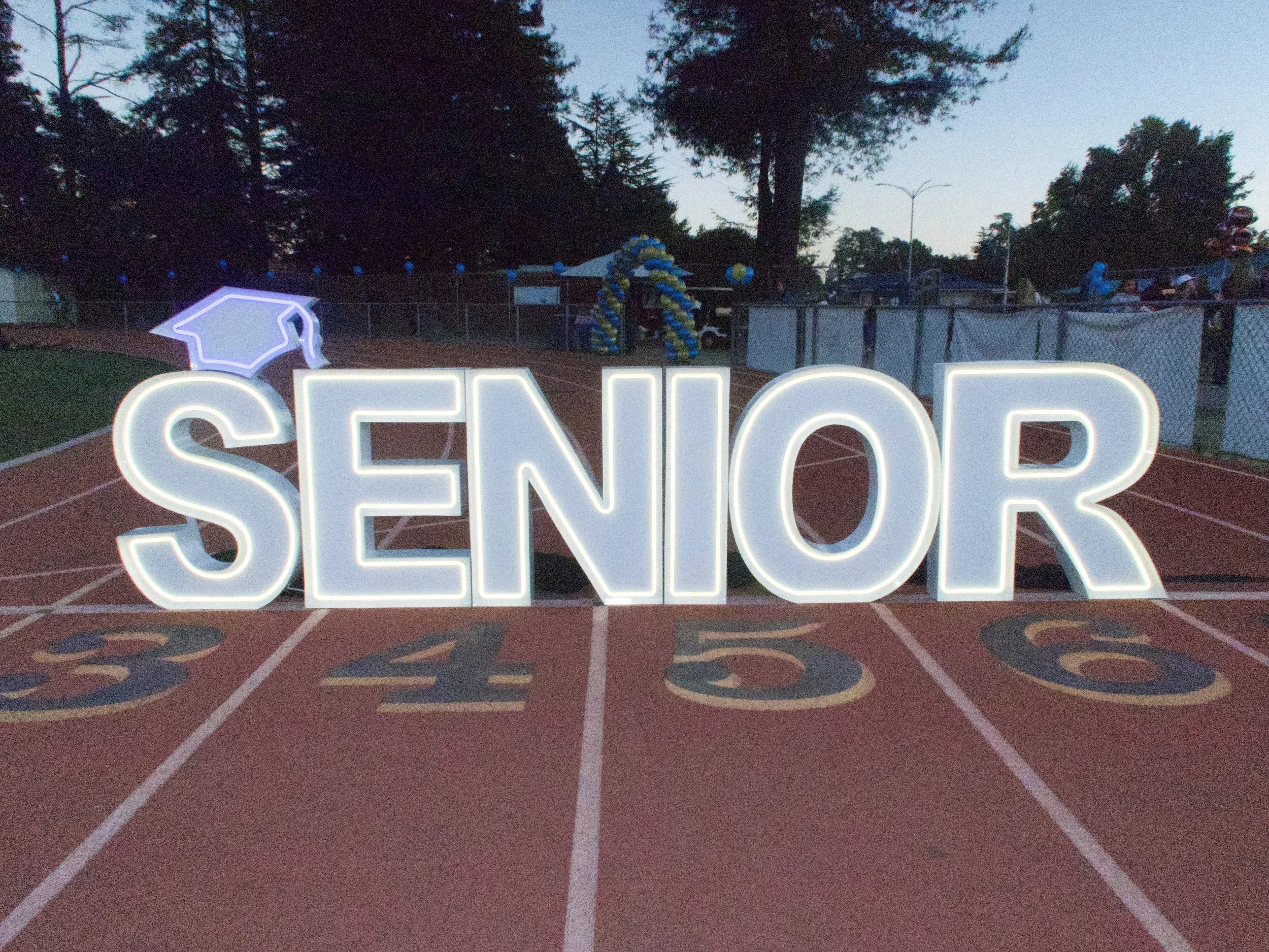 Large illuminated sign spelling 'SENIOR' with a graduation cap icon, placed on a numbered running track at dusk, with trees and balloons in the background.