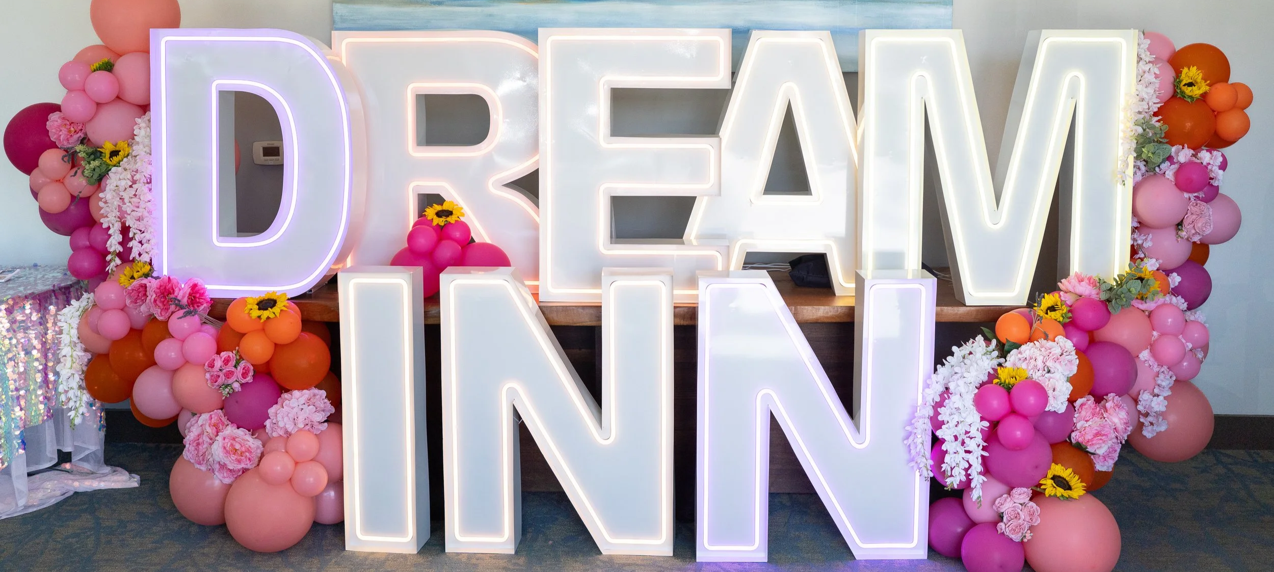 Large illuminated sign spelling 'DREAM INN' surrounded by pink, orange, and purple balloons and floral arrangements with sunflowers and pink roses at an event.