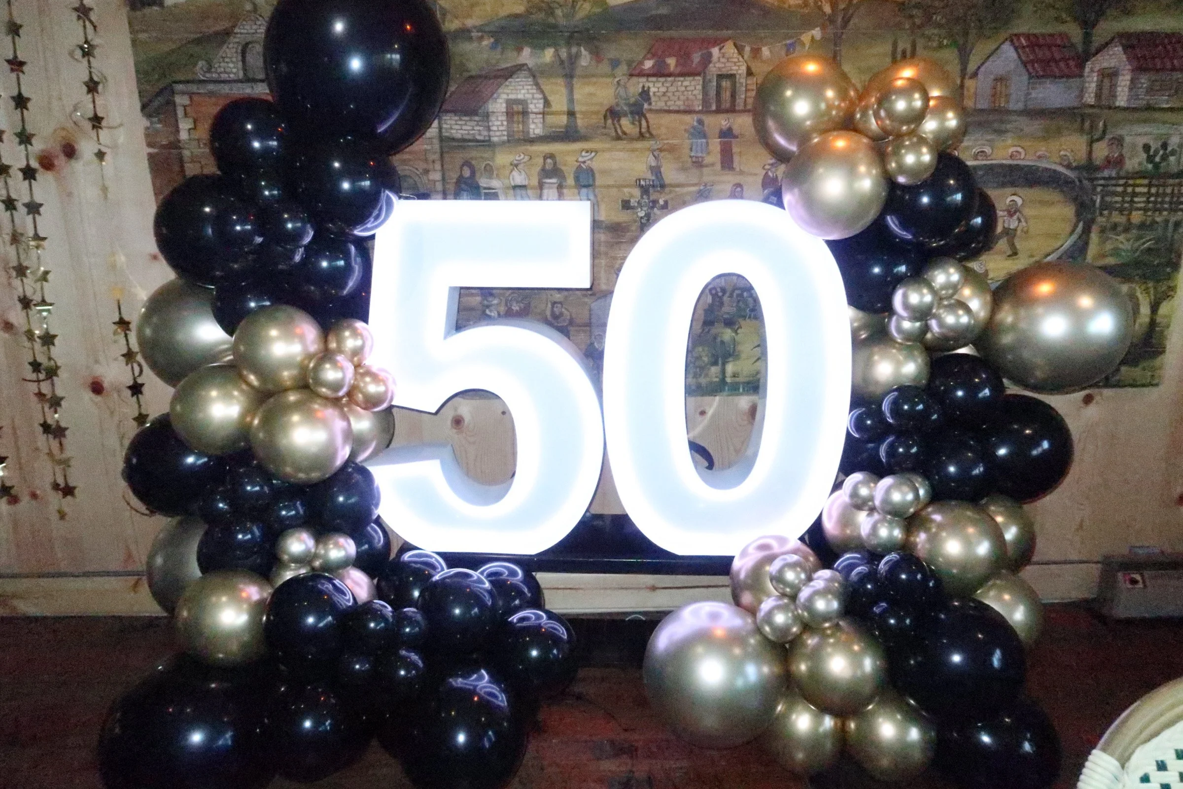 Decorative display celebrating a 50th anniversary or milestone with large illuminated number 50 surrounded by cluster of black, gold, and pearl-colored balloons.