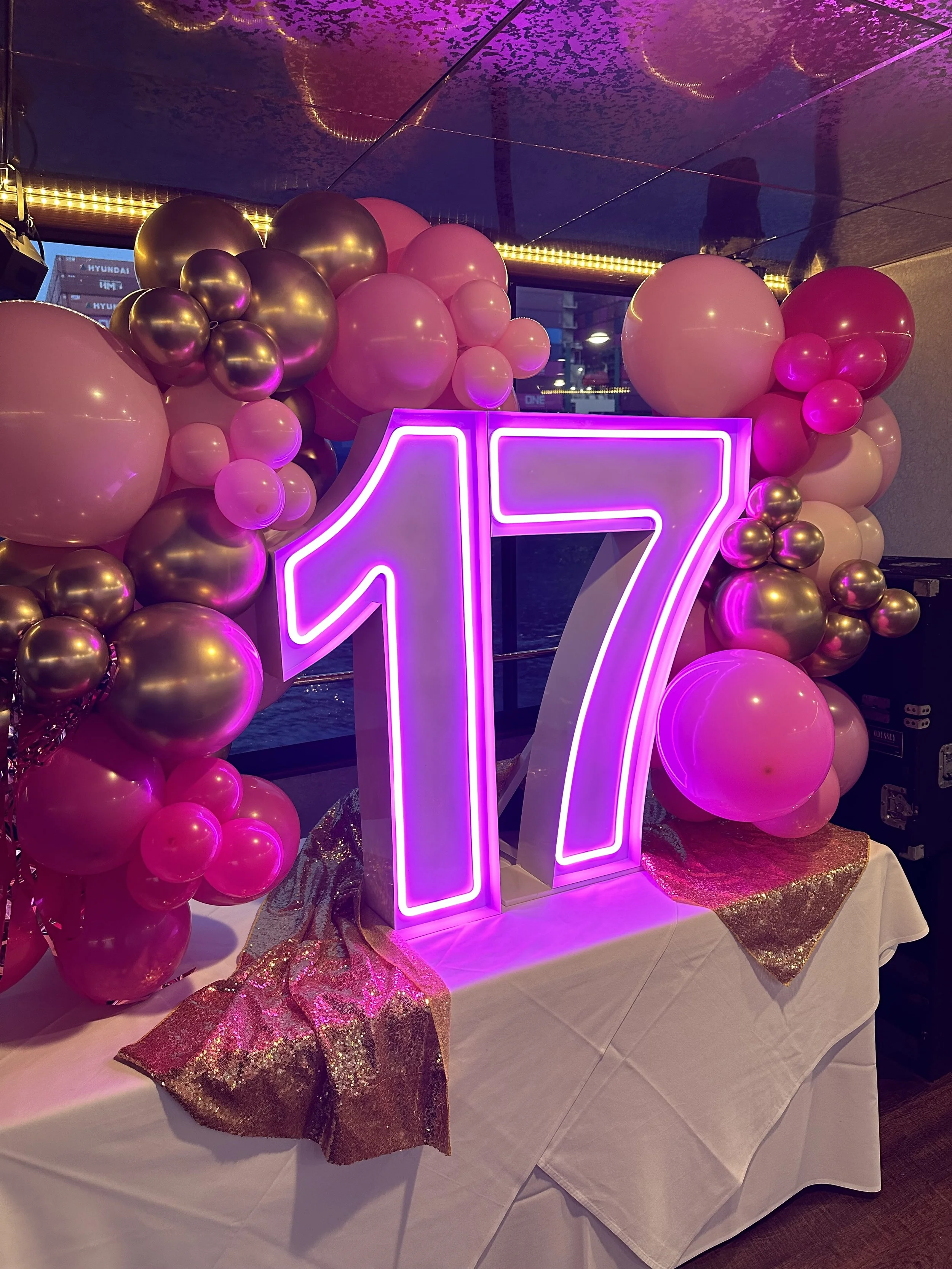 Large illuminated pink and purple neon number 17 sign surrounded by pink and gold balloons, on a table with a white tablecloth and gold glittery accents, celebrating a 17th birthday.