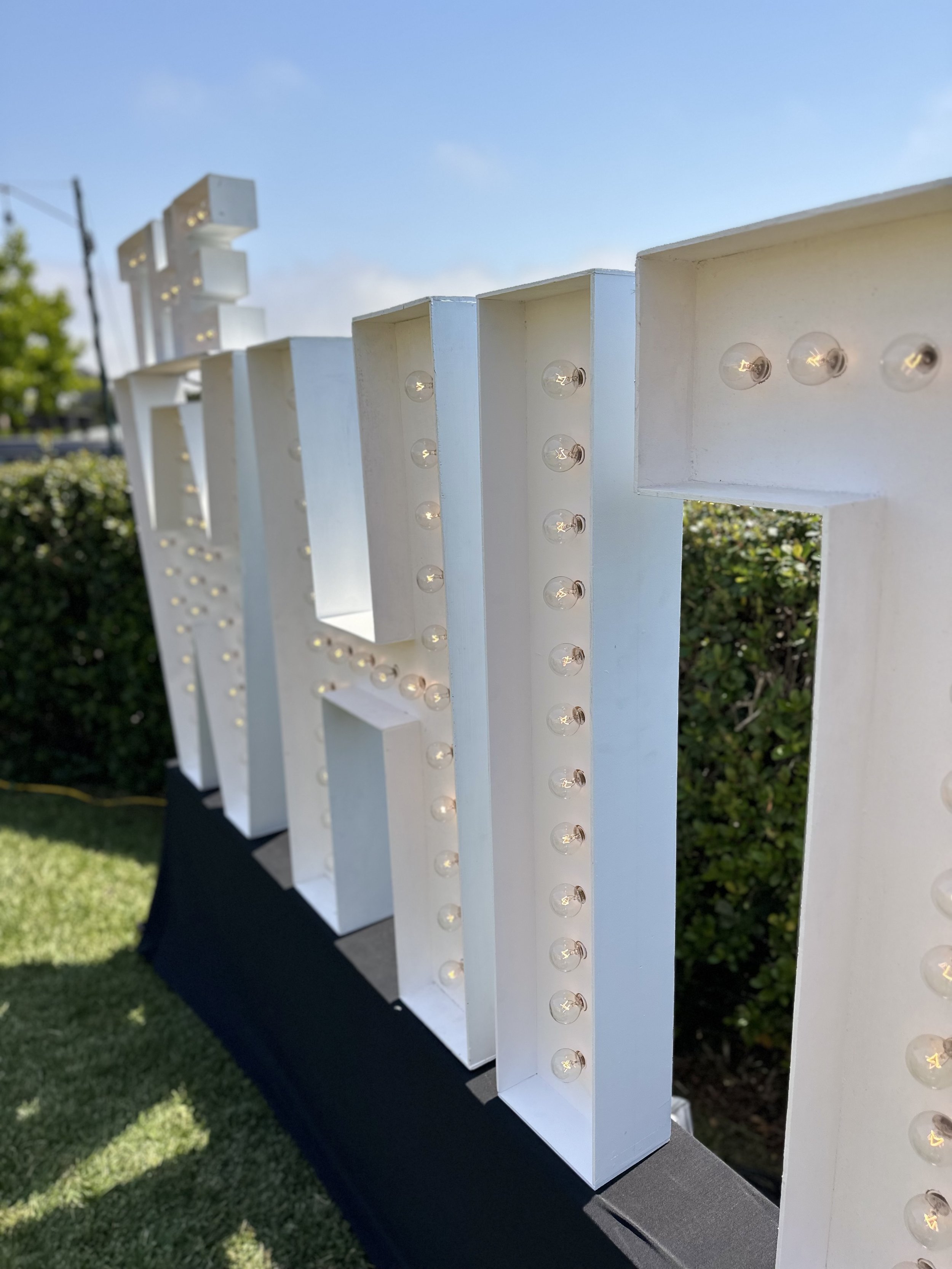White marquee letter with small light bulbs along the edges, outdoors on grass and bushes, under a blue sky.