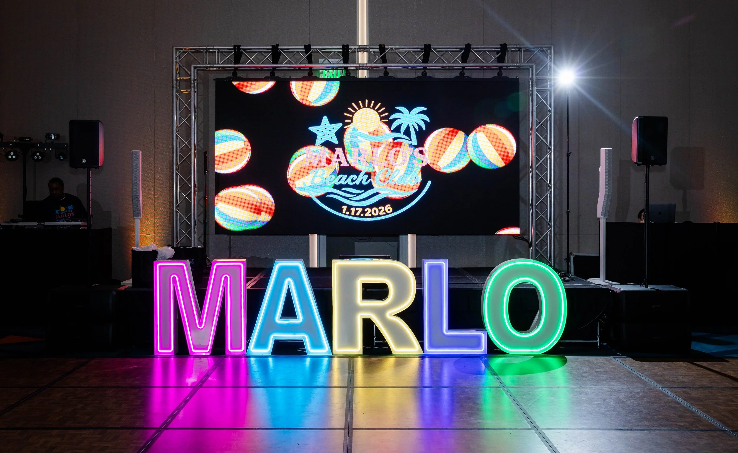 Colorful neon letters spelling 'MARLO' on the dance floor at a beach-themed event with a large digital screen displaying a tropical design and the date 1.17.2026.